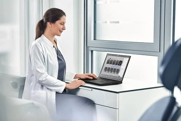 A female medical professional in a white coat working on a laptop at a windowed desk.