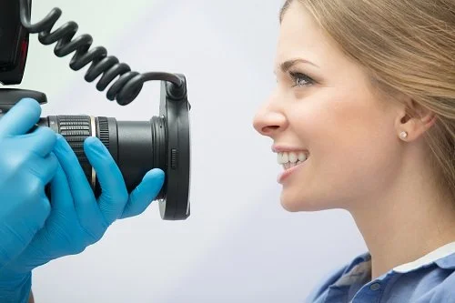A woman smiling as a person in blue gloves holds a camera close to her face for a photograph.