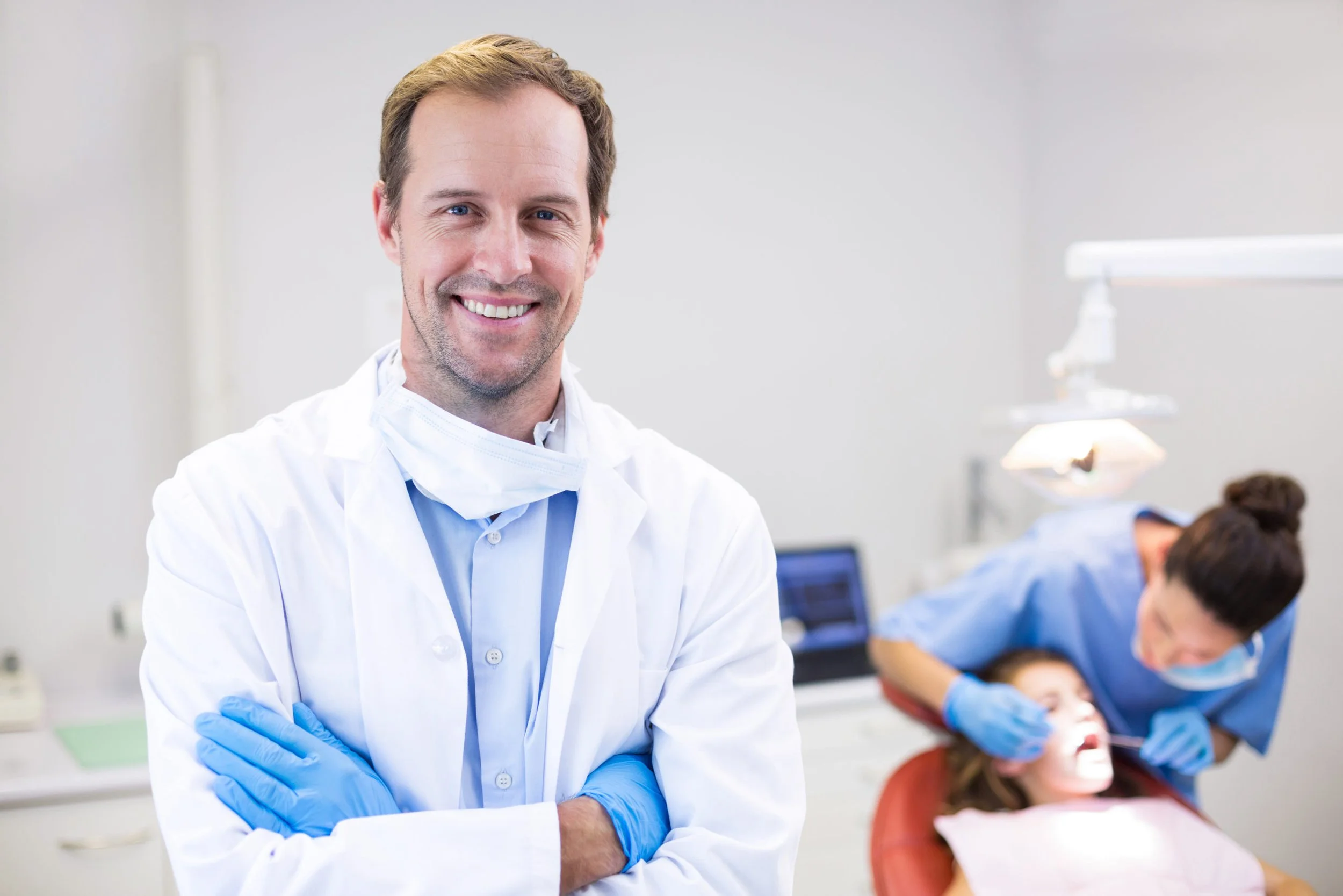 A male dentist in a white coat and blue gloves smiles at the camera in a dental clinic, with a female dental assistant attending to a young patient in the background.