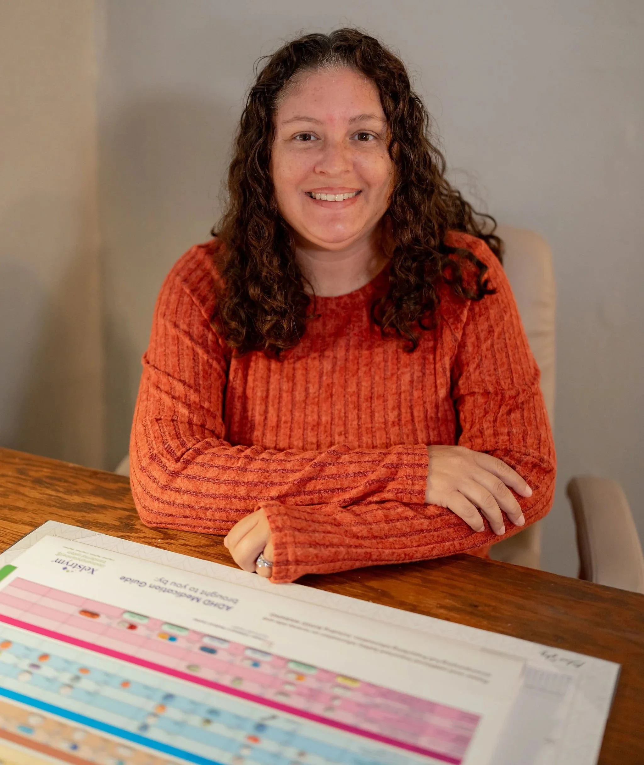 A woman with curly hair wearing an orange sweater, sitting at a wooden table with a colorful chart or poster in front of her.