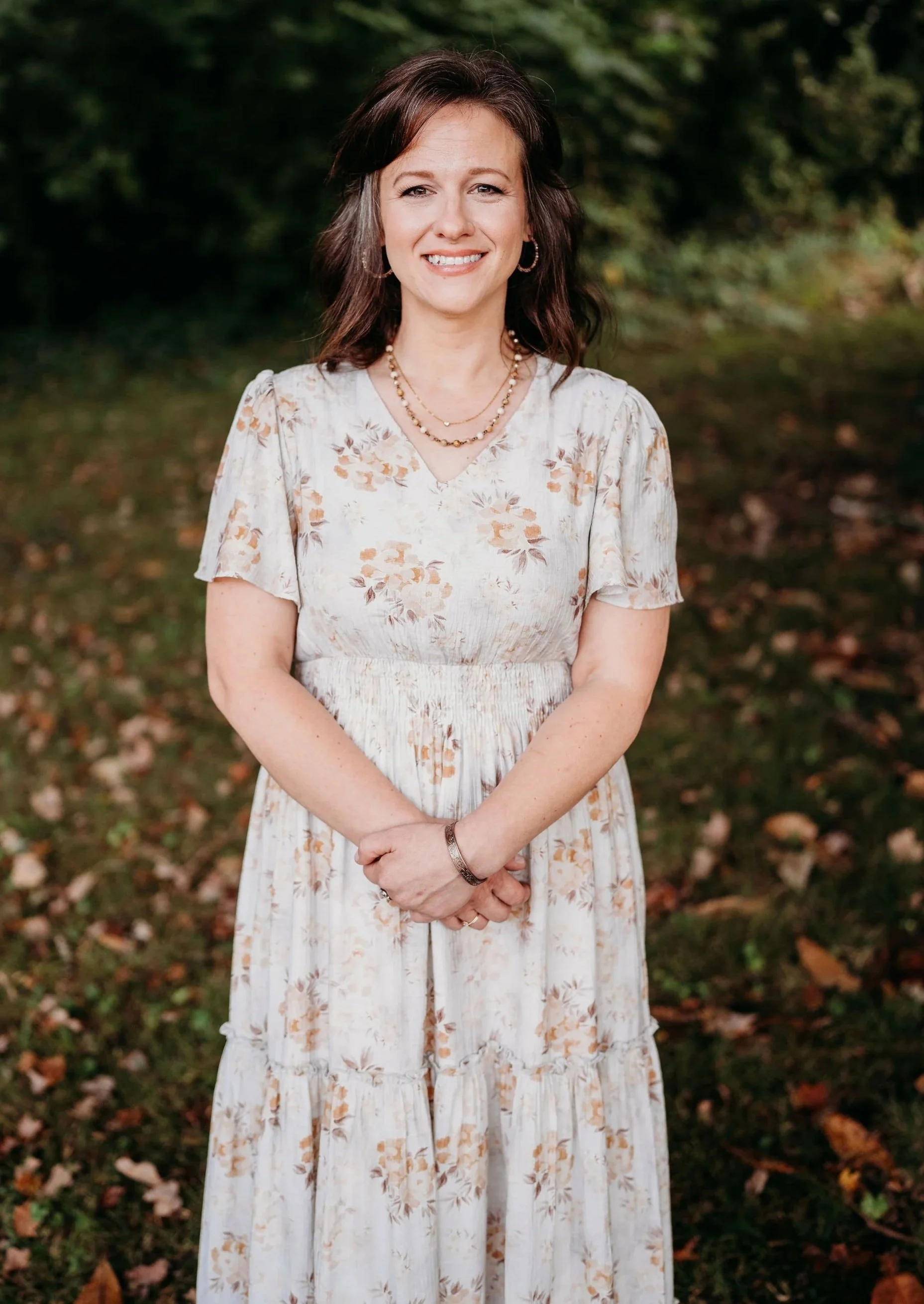A woman with shoulder-length dark hair, wearing a floral dress, standing outdoors on a grassy area with fallen leaves and green bushes in the background. She is smiling and looking at the camera.