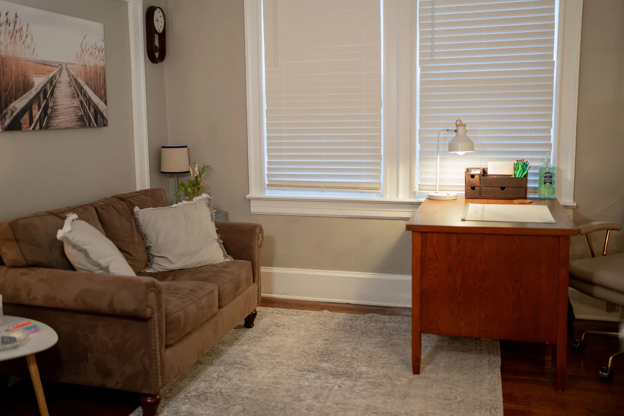Living room with beige sofa, pillows, window, wooden desk with lamp, office chair, and artwork on the wall.