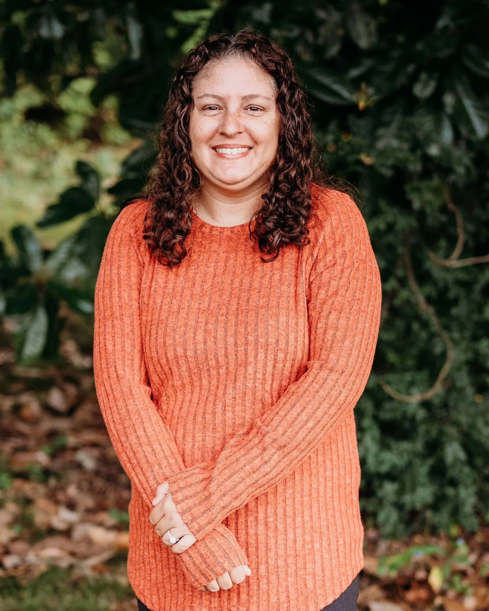 A woman with curly brown hair smiling, standing outdoors in front of green foliage, wearing an orange long-sleeve sweater.
