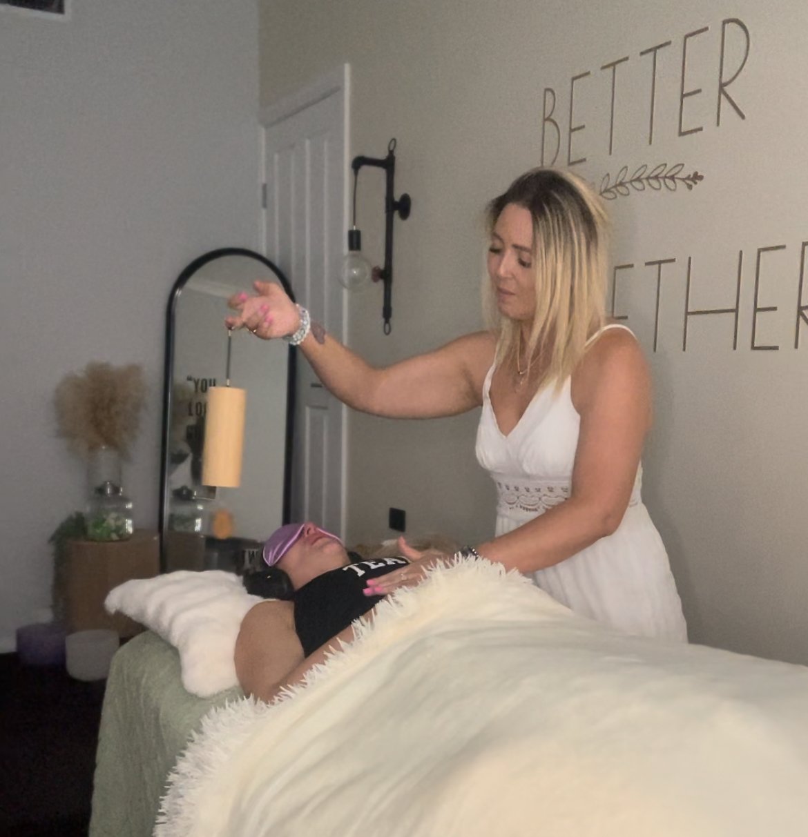 A woman in a white dress performing a relaxation or wellness therapy session on a person lying on a table, covered with a blanket and wearing an eye mask. The therapist is holding a sound healing instrument. The words 'Better Together' are on the wal