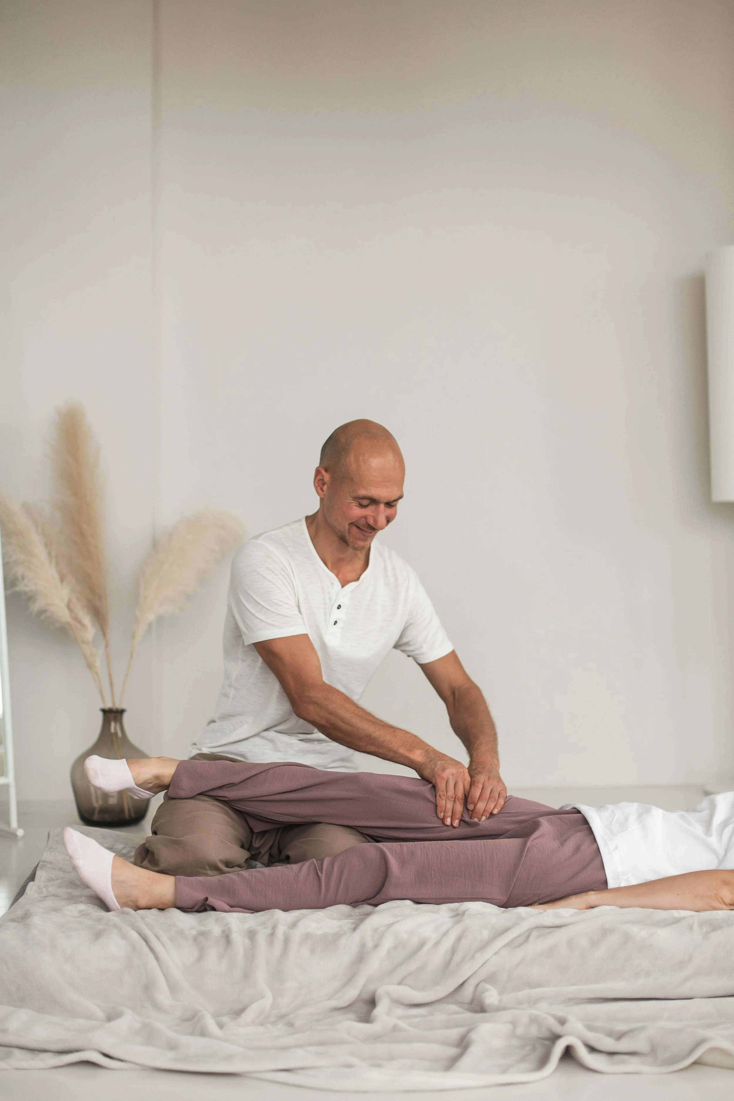 A person receiving a massage from a therapist in a spa setting with neutral decor, including pampas grass in a vase, on a massage table covered with a beige sheet.