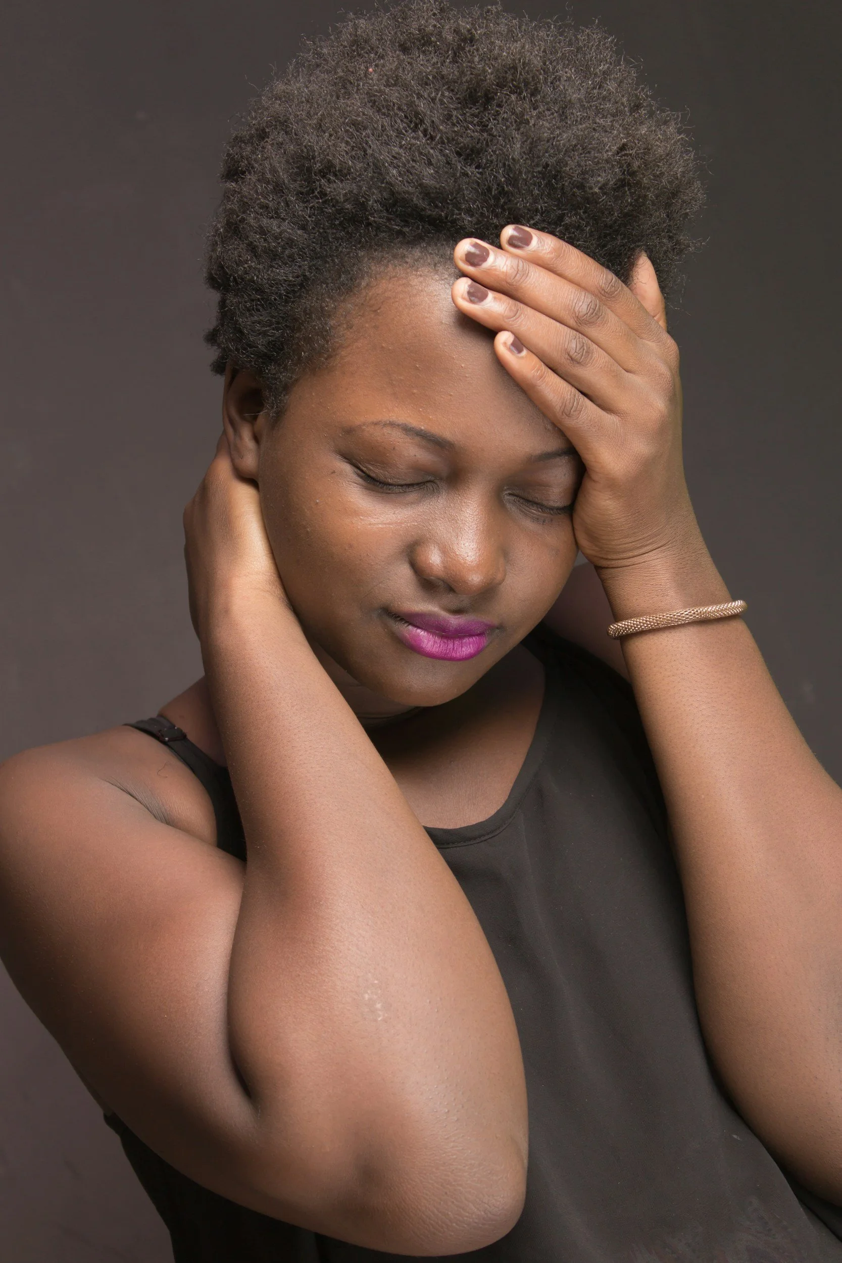 A woman with short, curly black hair, holding her forehead with one hand and her neck with the other, wearing a black tank top and a gold bracelet, with her eyes closed and a calm, contemplative expression.