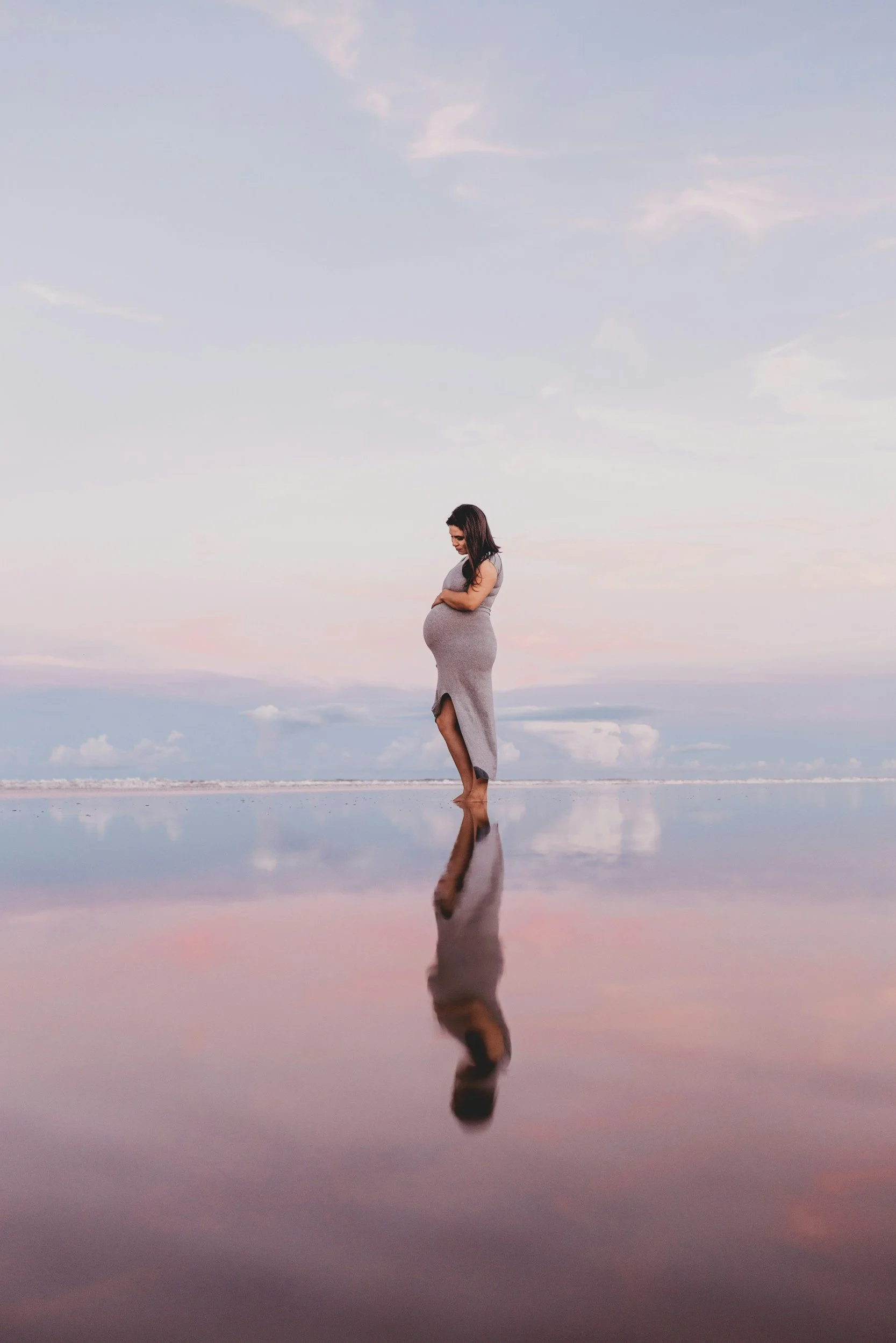 A pregnant woman standing barefoot on a reflective wet surface at sunset, looking down at her belly, with a soft sky background and clouds.