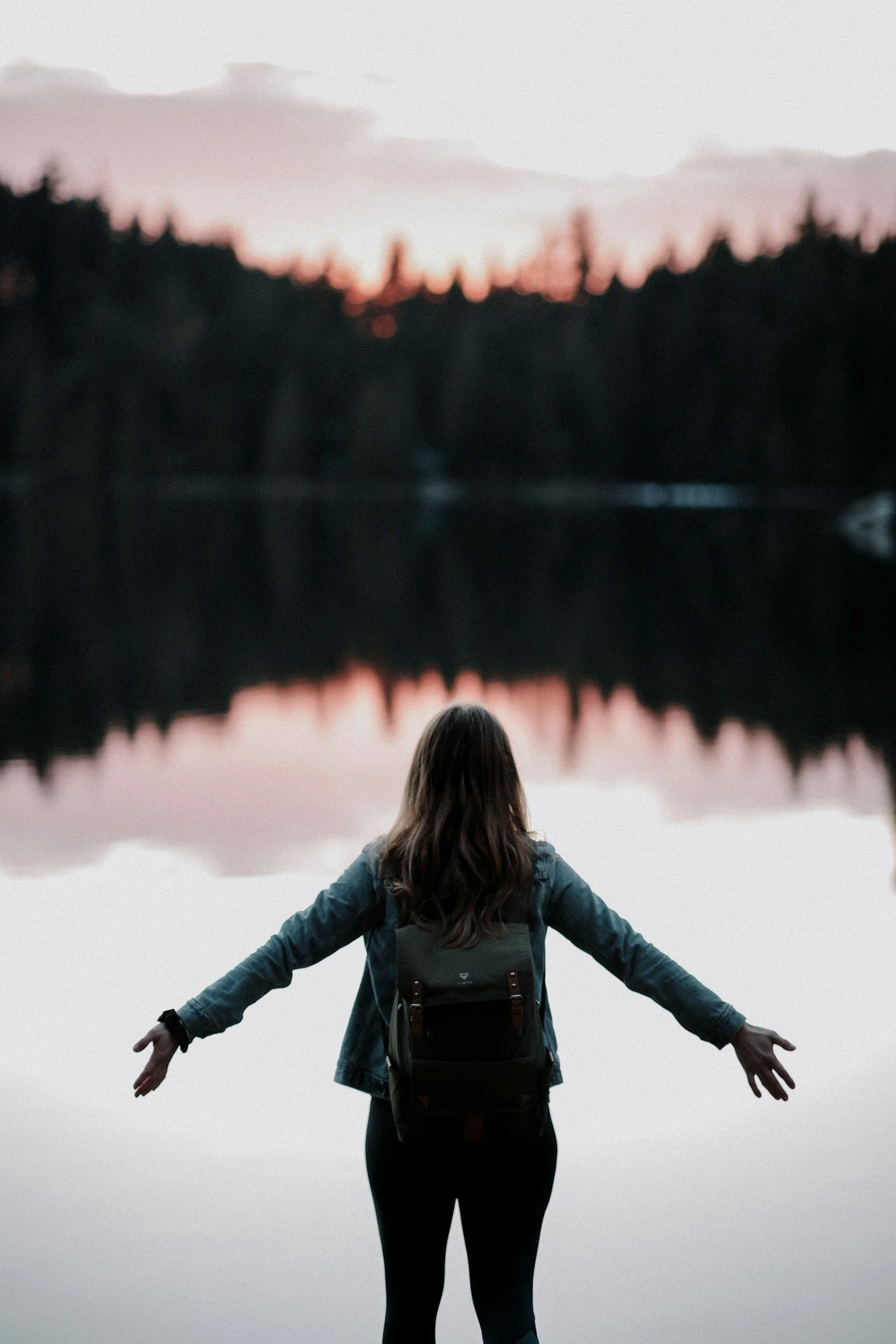 A woman with long hair, wearing a denim jacket and a backpack, standing on a dock with her arms outstretched, facing a calm lake reflecting a silhouette of trees and a pink-tinged sky during sunset.