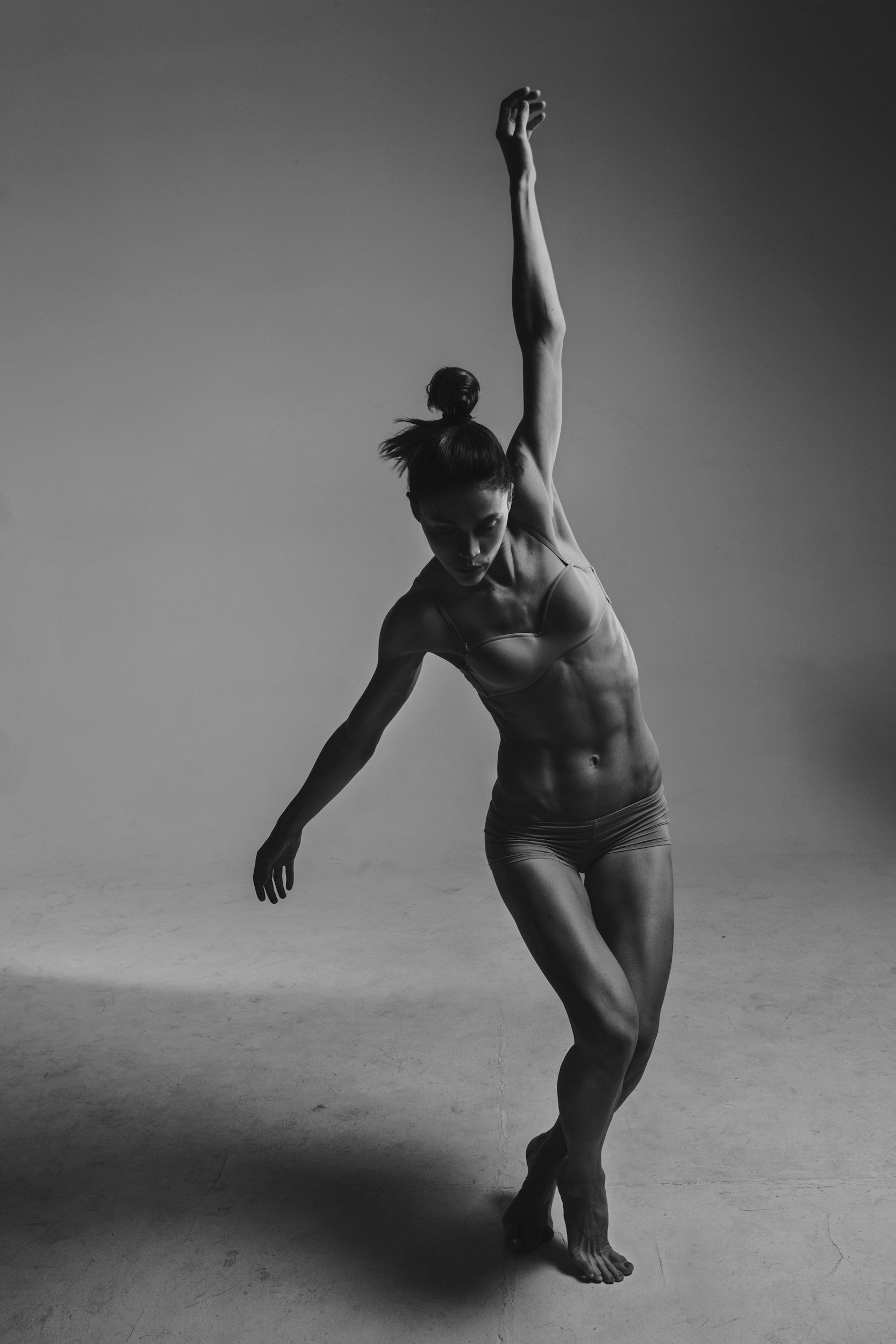 Black and white photo of a female dancer with an athletic build, wearing a light-colored sports bra and shorts, striking a dynamic pose with one arm raised above her head and the other extended outward, against a plain floor and wall background.