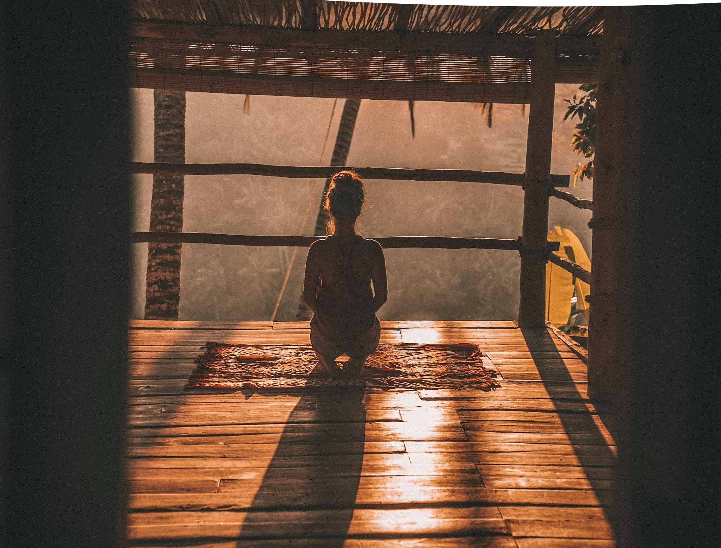 A person sitting on a wooden floor near a railing, looking out at a sunset over a mountain landscape with trees and power lines, framed by the structure of a thatched roof and wooden beams.