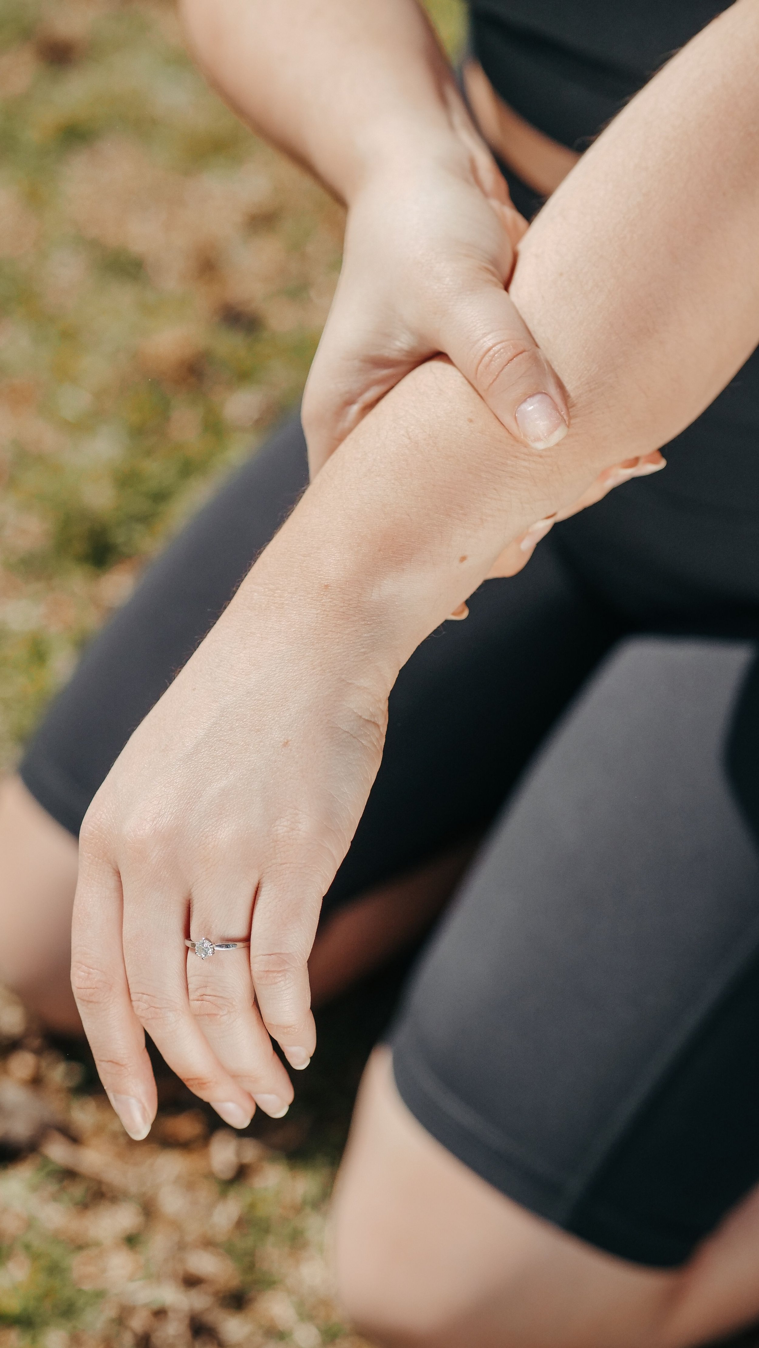 Close-up of a woman’s hand with a diamond engagement ring, resting on her thigh, outdoors with blurred grass background.
