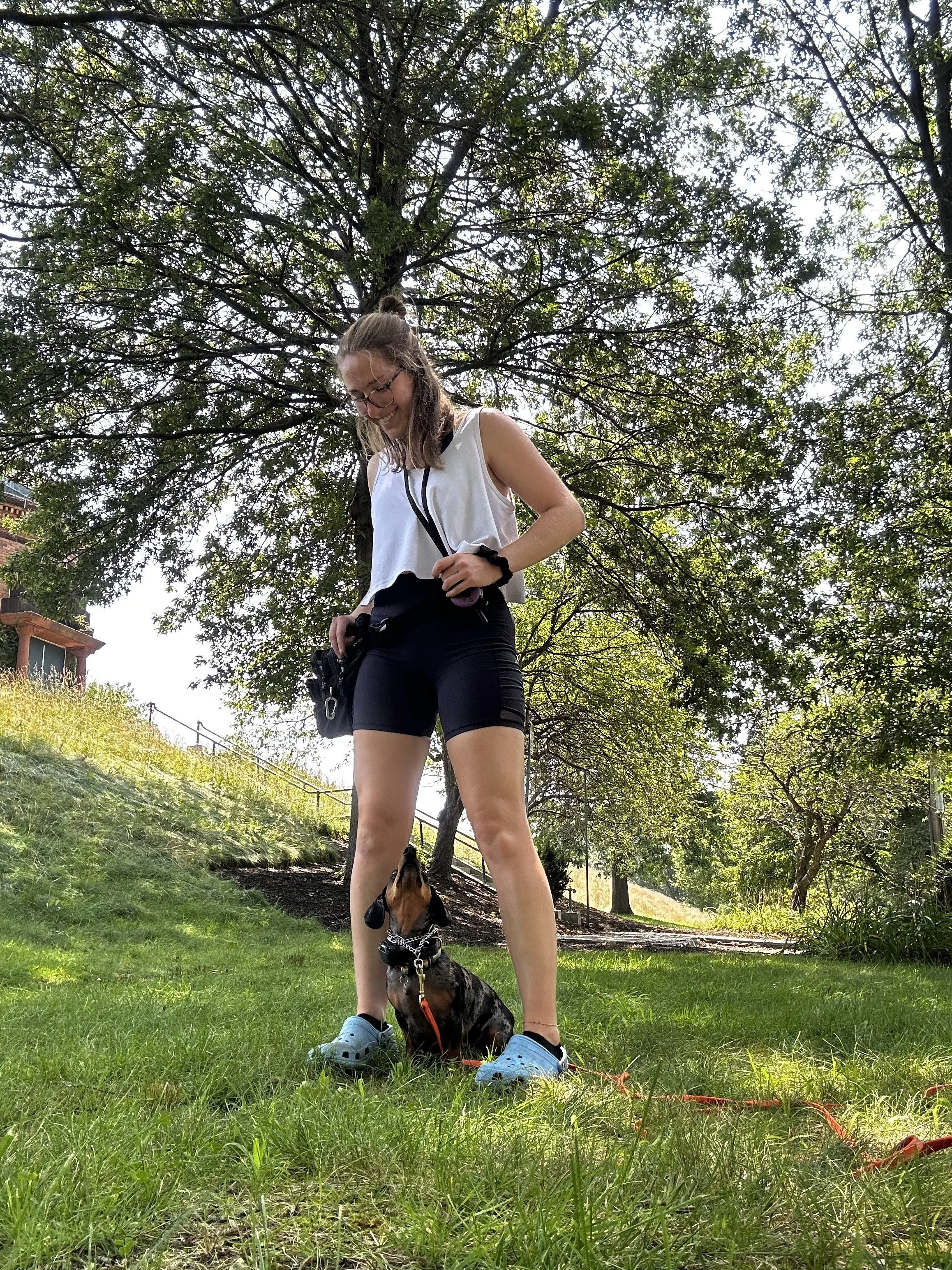 A young woman in athletic clothing standing on grass, looking down and smiling at a small dog sitting in front of her. The dog is wearing a collar and leash, and the scene is outdoors under a tree with green foliage.