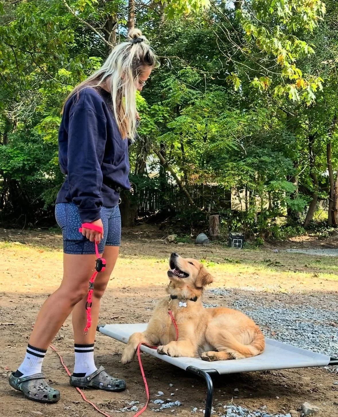 A woman stands next to a golden retriever puppy lying on a raised dog bed outdoors, surrounded by trees. The woman is wearing a navy sweatshirt, blue patterned shorts, white socks with black stripes, and crocs. She holds a pink leash attached to the puppy, who is smiling and looking up at her.