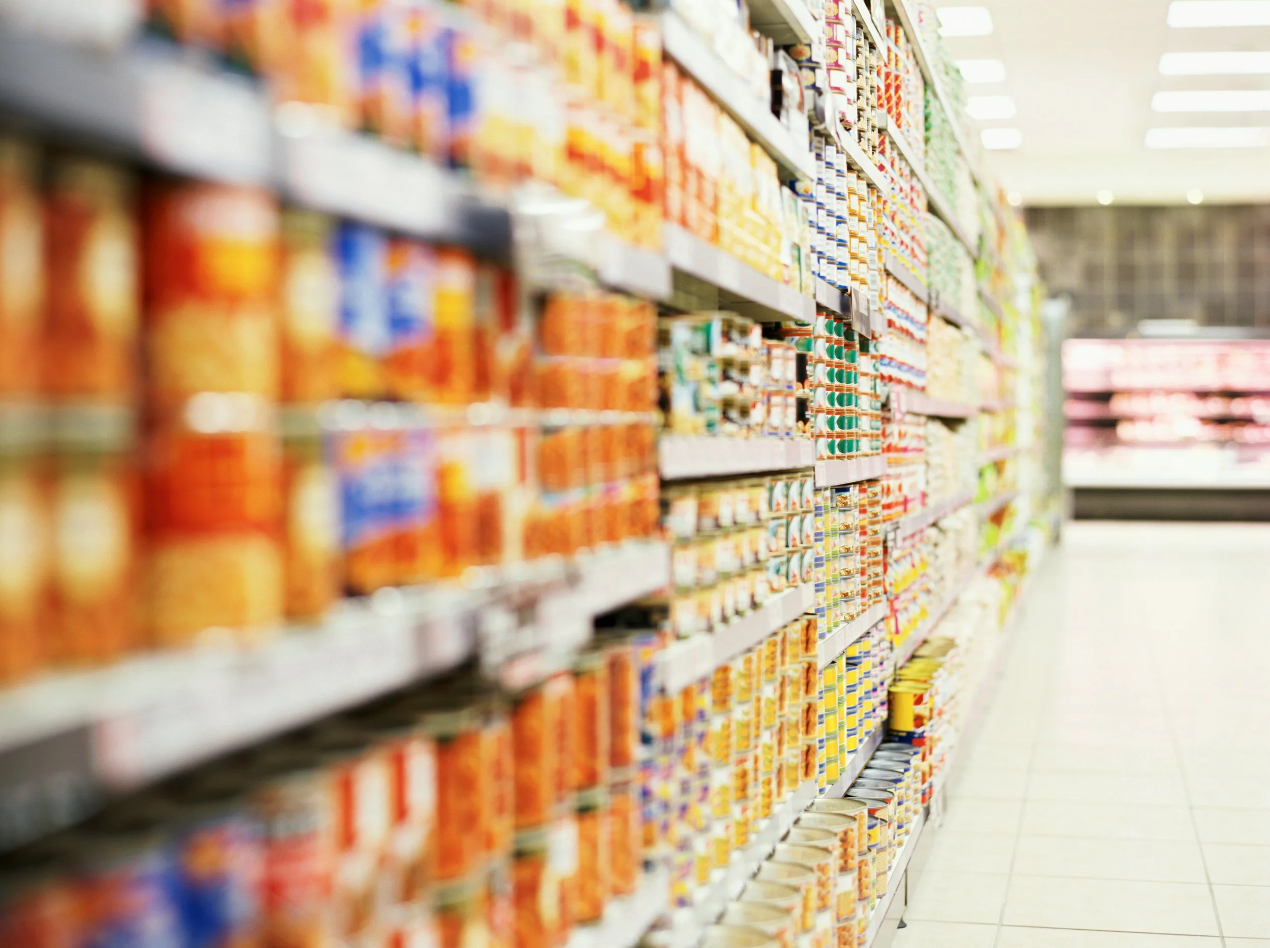 Aisle of shelves stocked with canned goods and packaged food products in a grocery store, with a tiled floor and bright lighting.