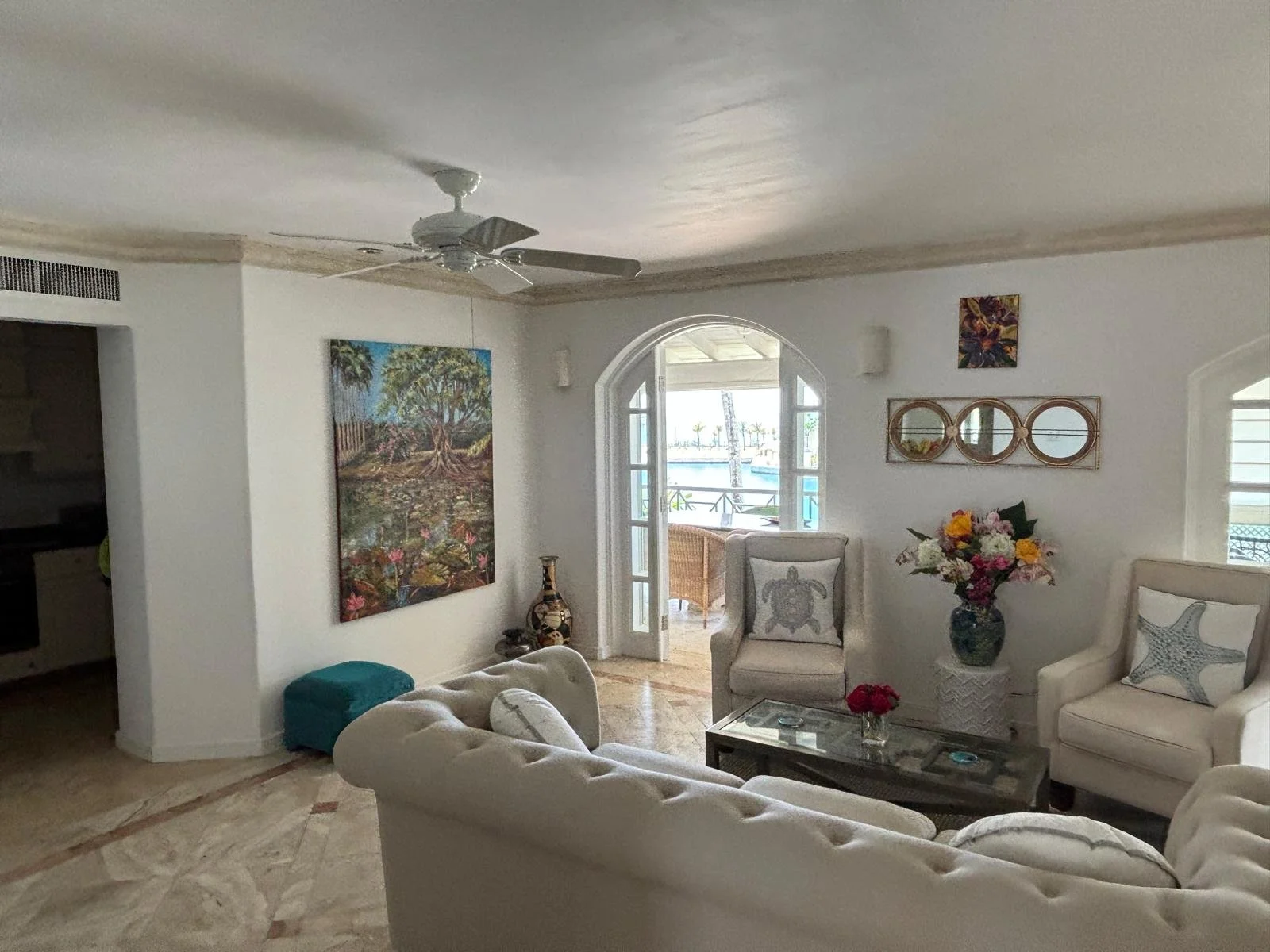 Bright living room with white walls, a ceiling fan, and a large window. Includes beige sofas, a glass coffee table, two upholstered armchairs with decorative starfish and turtle pillows, and a vase with flowers. Artwork and round mirrors adorn the walls.