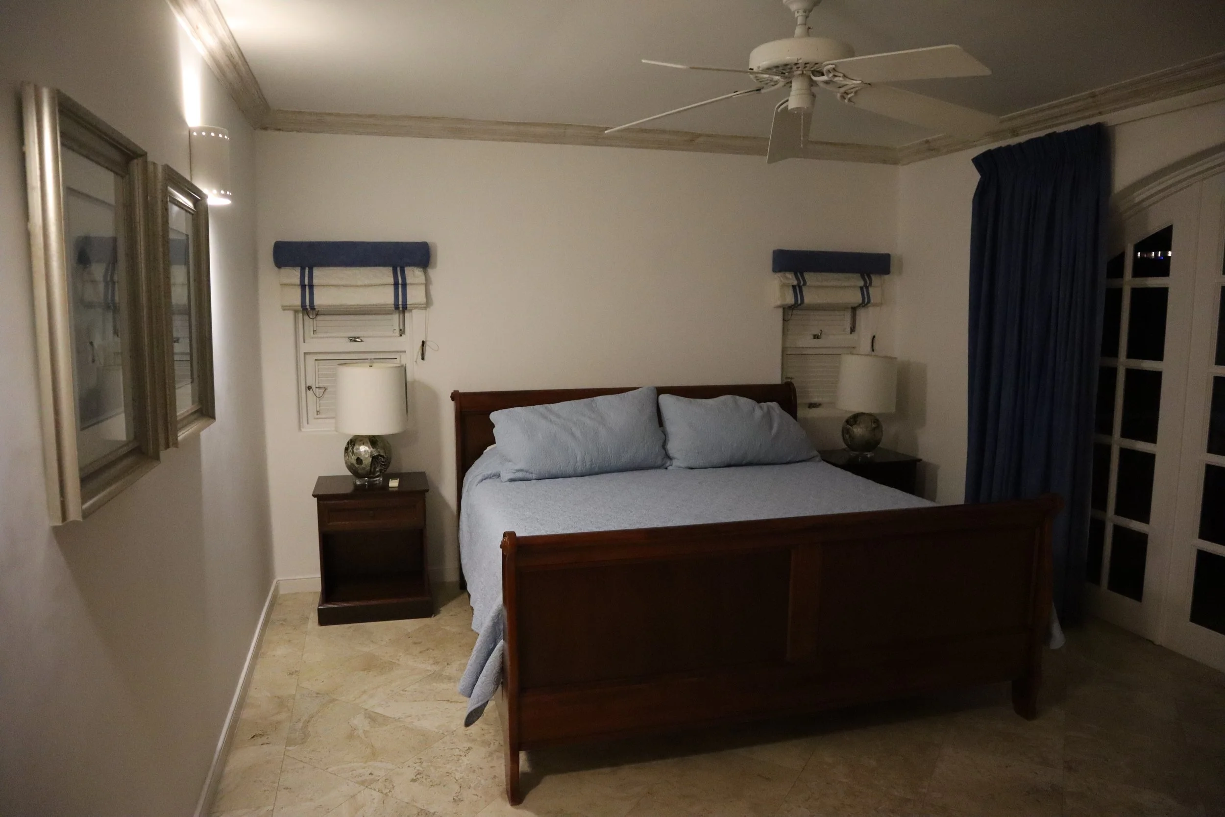 Bedroom with a wooden bed, blue bedspread, bedside tables with lamps, curtains, and framed artwork on the wall.