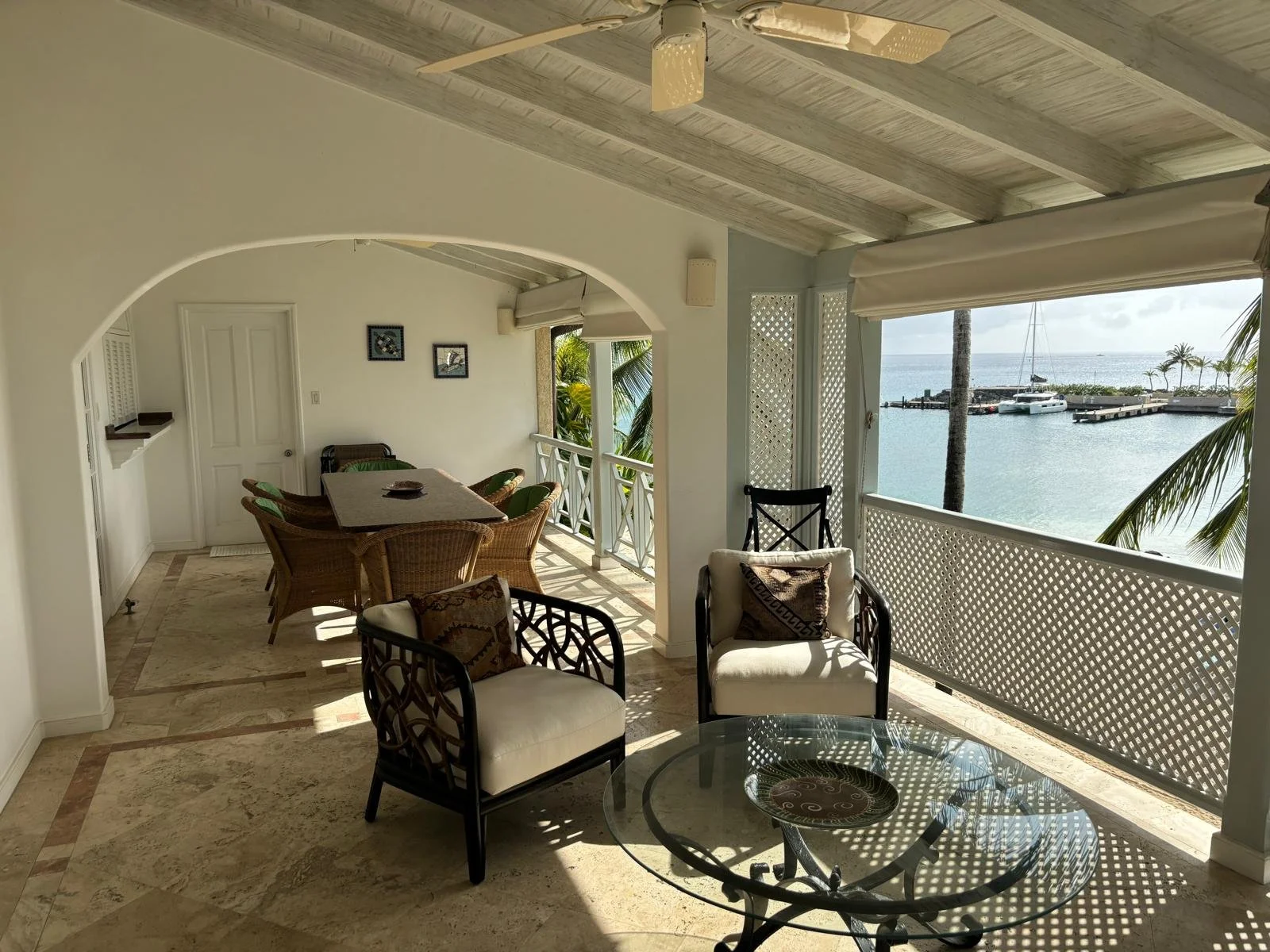 A covered balcony with a tiled floor features wicker chairs and a table, with additional cushioned chairs around a glass coffee table. The balcony overlooks a marina with boats and palm trees, offering a view of the ocean under a partly cloudy sky.