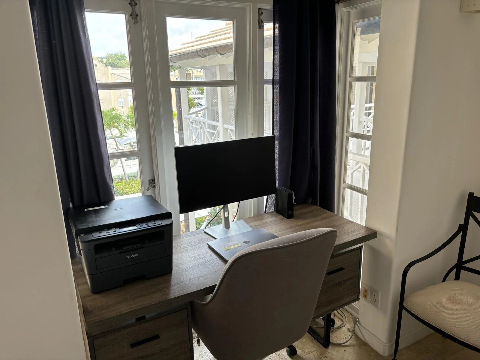 Home office setup with a printer, computer monitor, and laptop on a wooden desk. There's a cushioned chair in front and windows with dark curtains in the background.
