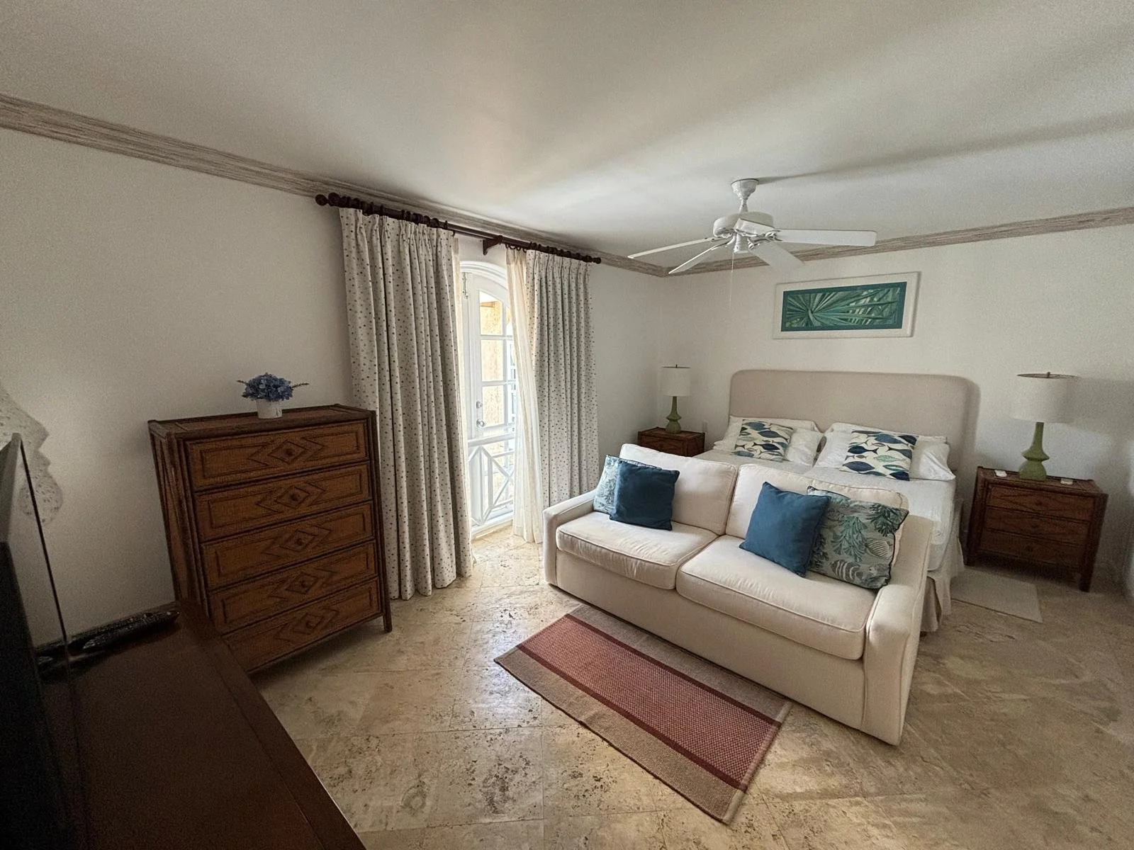 A bedroom with a bed, cream-colored sofa, and green leaf artwork. Wooden bedside tables with lamps sit on either side of the bed. A dresser with a small blue vase is near a window with patterned curtains. A ceiling fan is above.