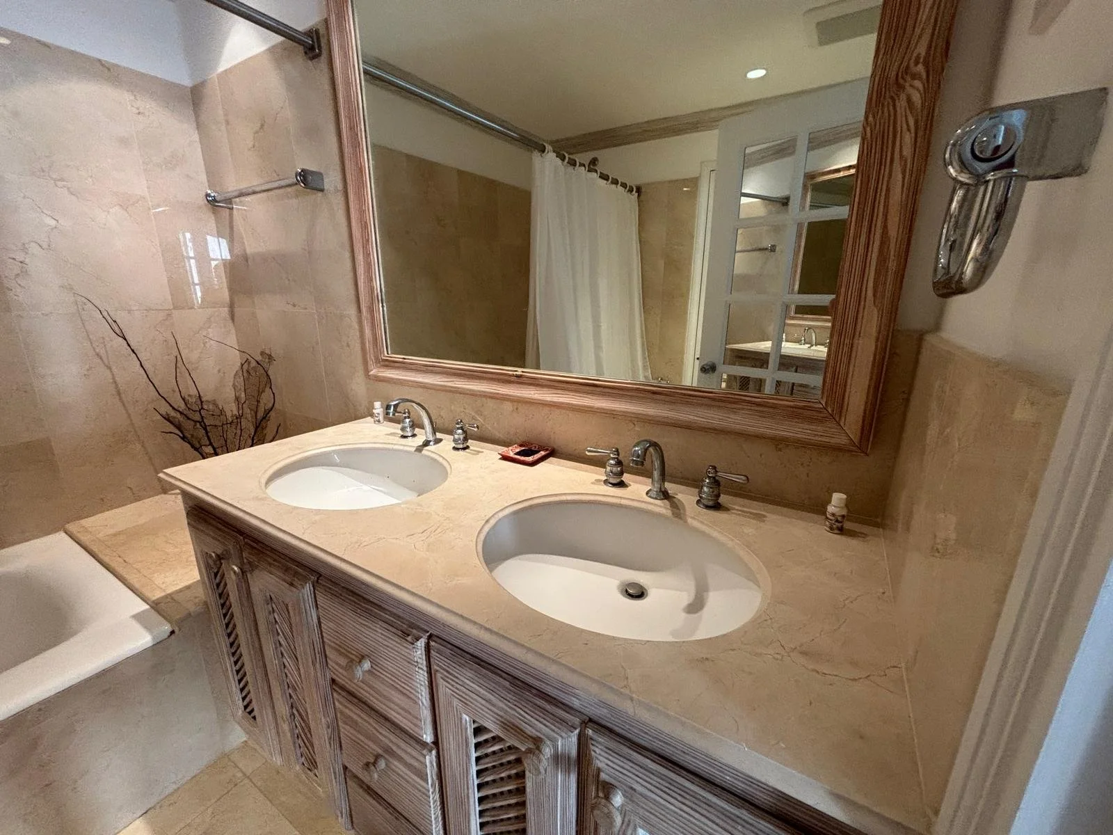 Modern bathroom with a marble countertop, dual sinks, large mirror, and beige tile walls. A bathtub and shower curtain are visible.