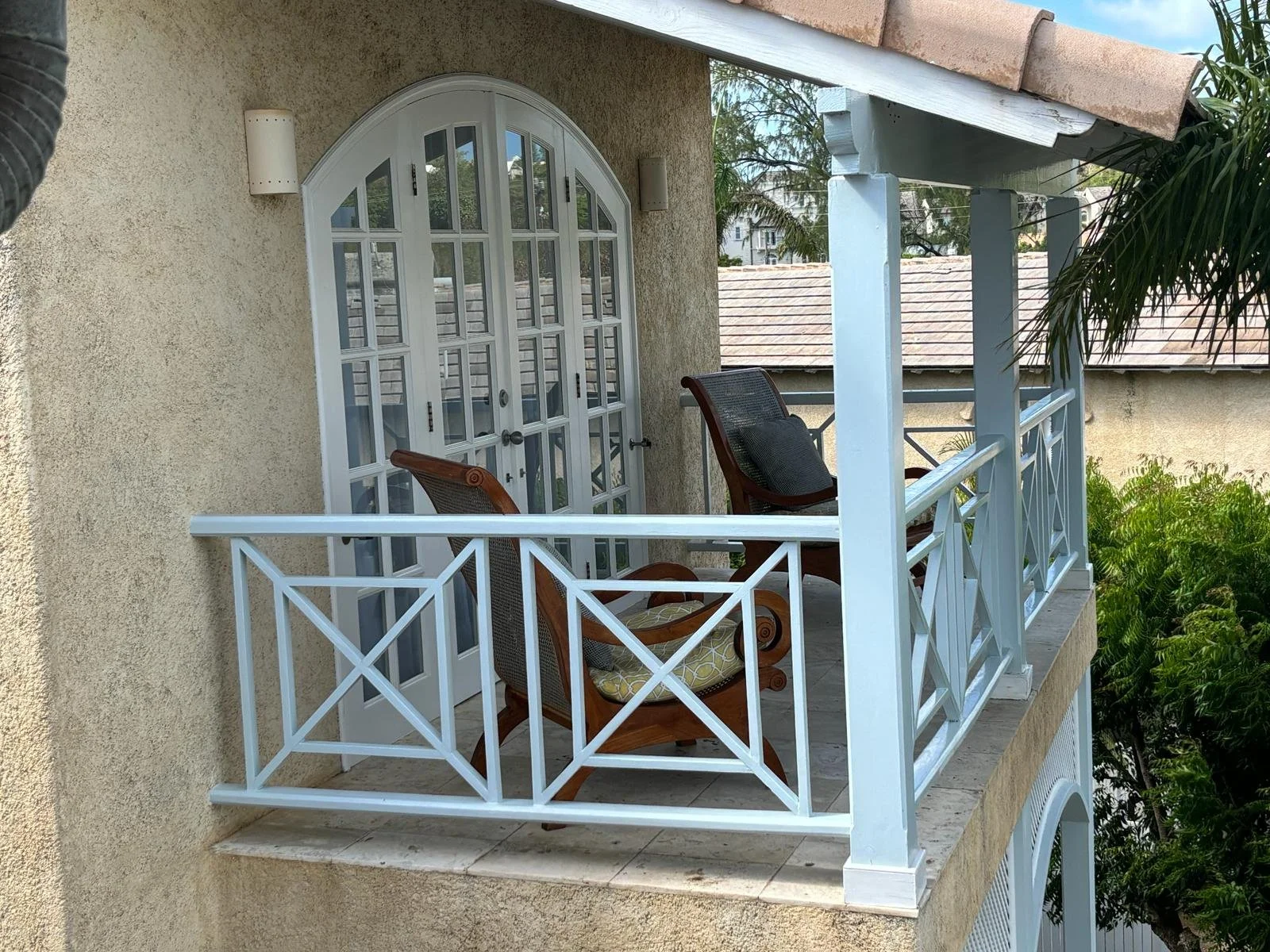 A small balcony with a wooden rocking chair and a cushion, enclosed by a light blue railing, next to an arched glass door on a stucco building.