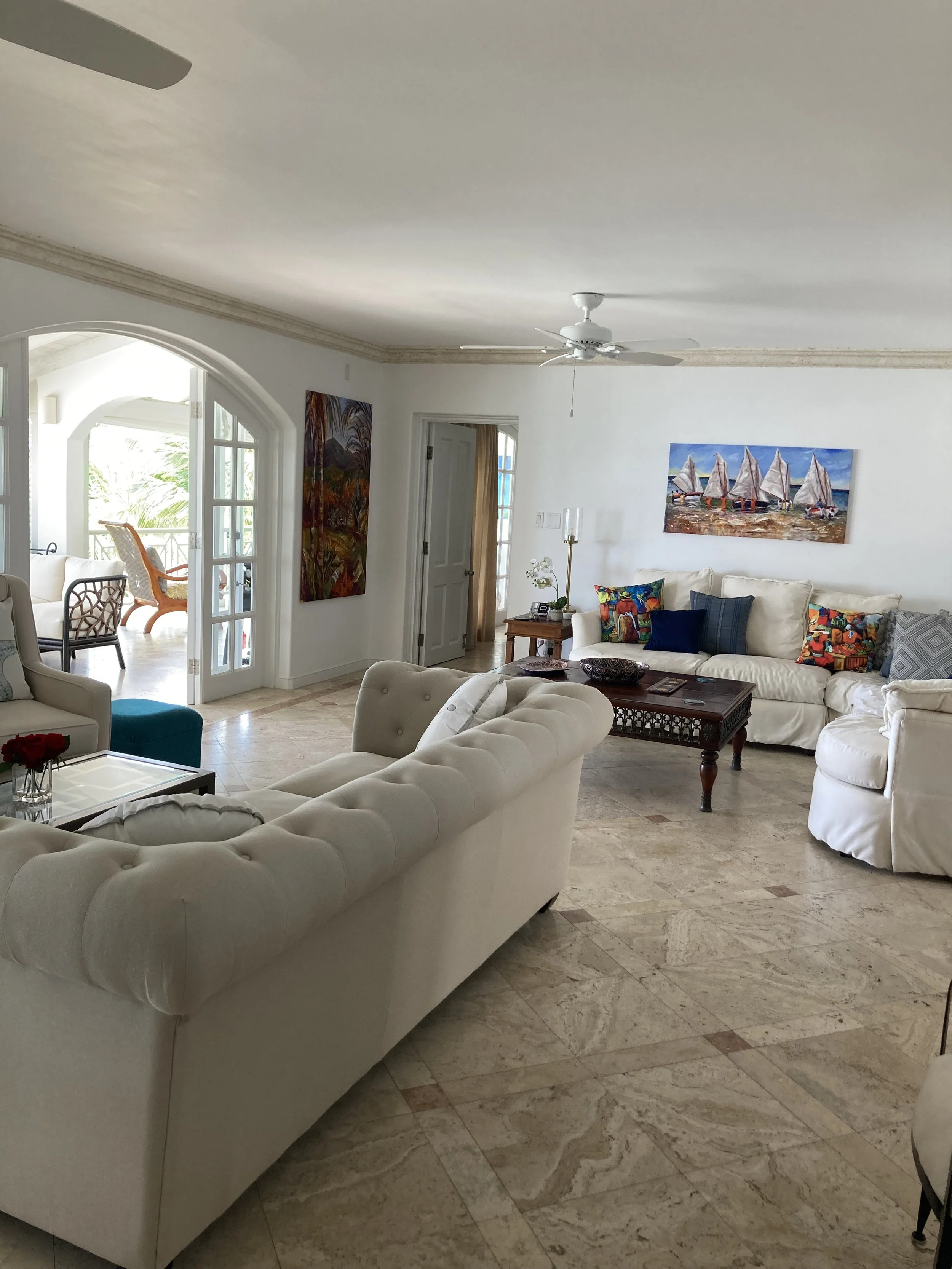 Elegant living room with white sofas and a marble floor, featuring paintings on the walls and decorative pillows. A ceiling fan and sliding glass doors leading to a patio are visible.