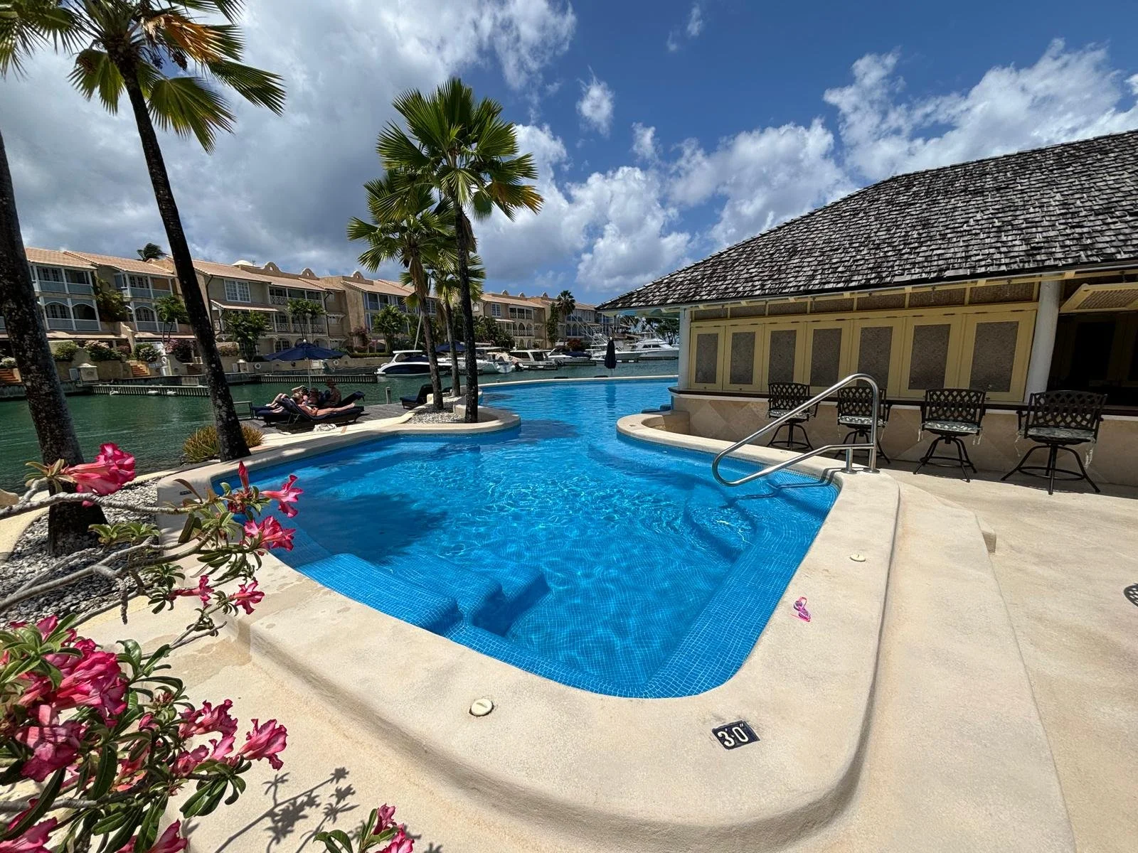 Tropical resort poolside area with palm trees, pink flowers, and lounge chairs next to a canal with yachts and waterfront buildings.