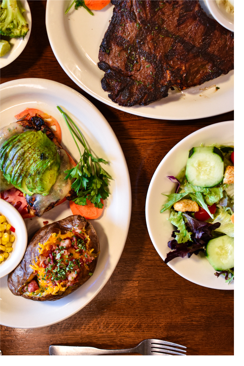 A table with several plates, including salads, broccoli, t-bone steak, and chicken bellaire.