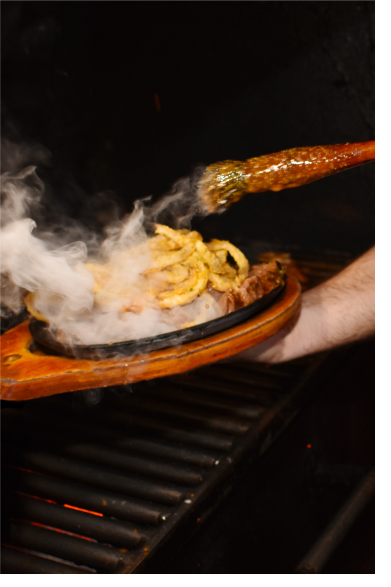 A man holds a fajita plate with the sizzling steak for two, adding butter while smoke pours from the plate.