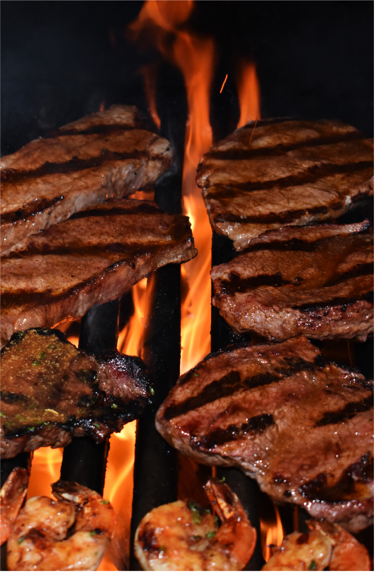 Several steaks and shrimp on the wood fire grill, with the flames coming up through the bars