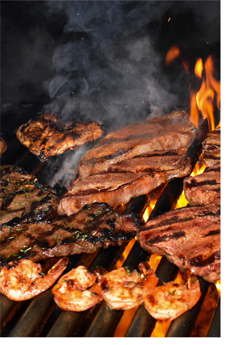 Image of steaks, chicken, and shrimp on the wood fire grill, with dramatic smoke, and flames coming up through the bars.