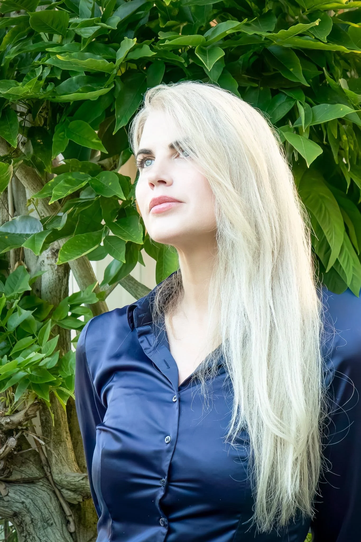 A woman with long, platinum blonde hair wearing a dark navy blue satin shirt, standing outdoors near green foliage and a tree.