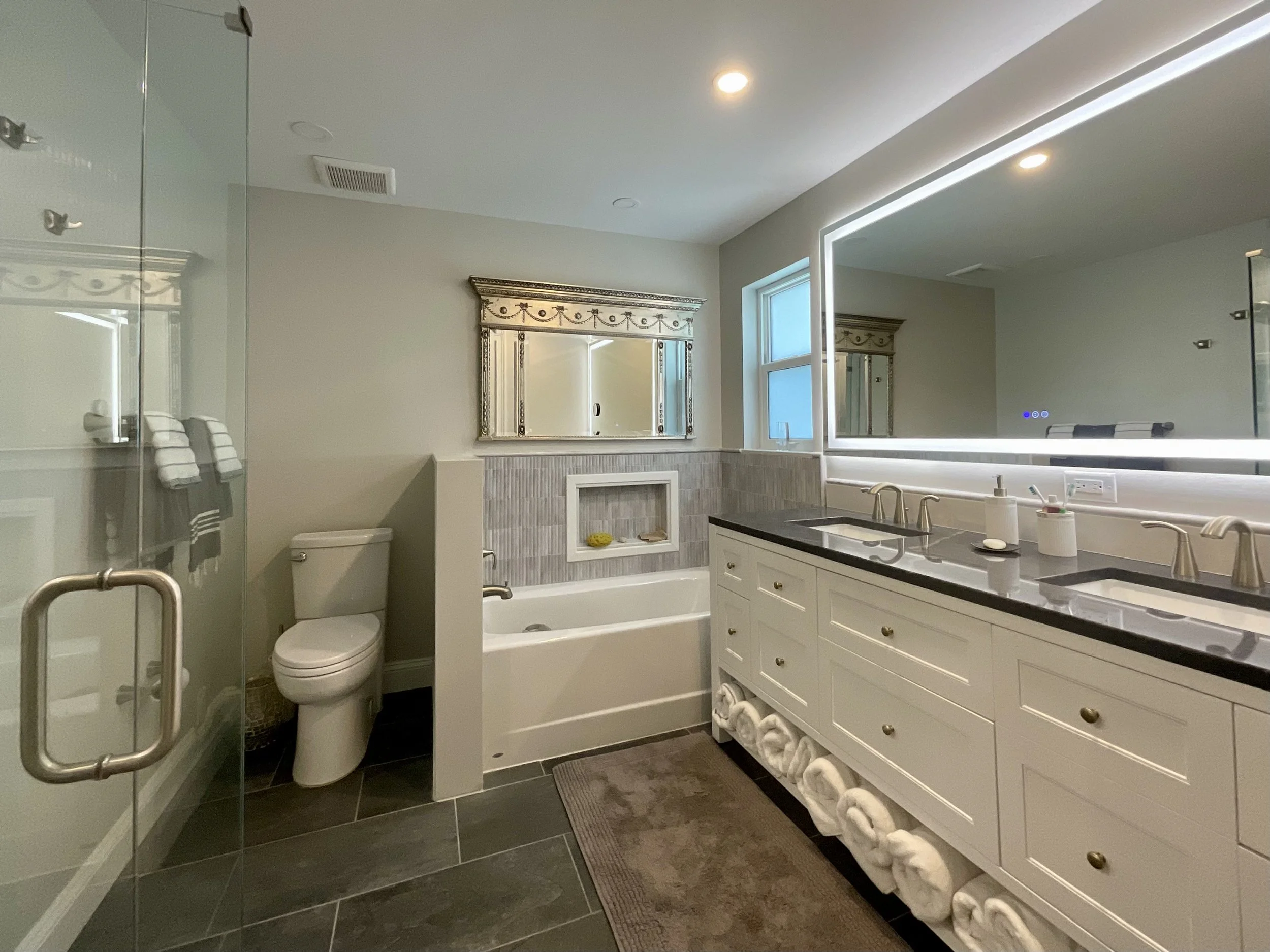 Master bathroom with double vanity, large illuminated mirror, bathtub with small window, half wall for toilet privacy, and glass-door shower.