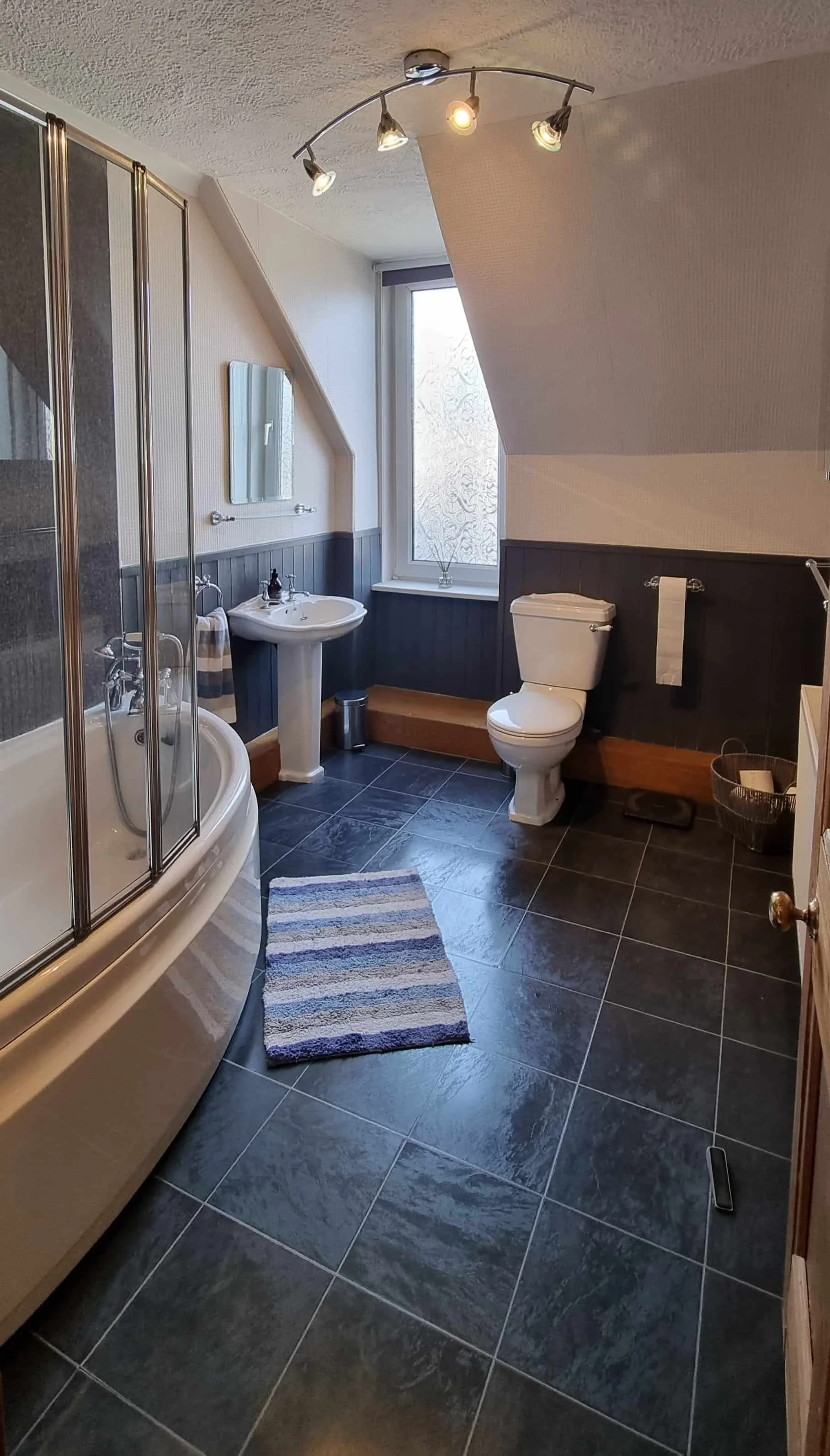 A bathroom with a black tile floor, a shower with a curved glass door, a pedestal sink, a toilet, a window with frosted glass, and a striped rug in front of the shower.