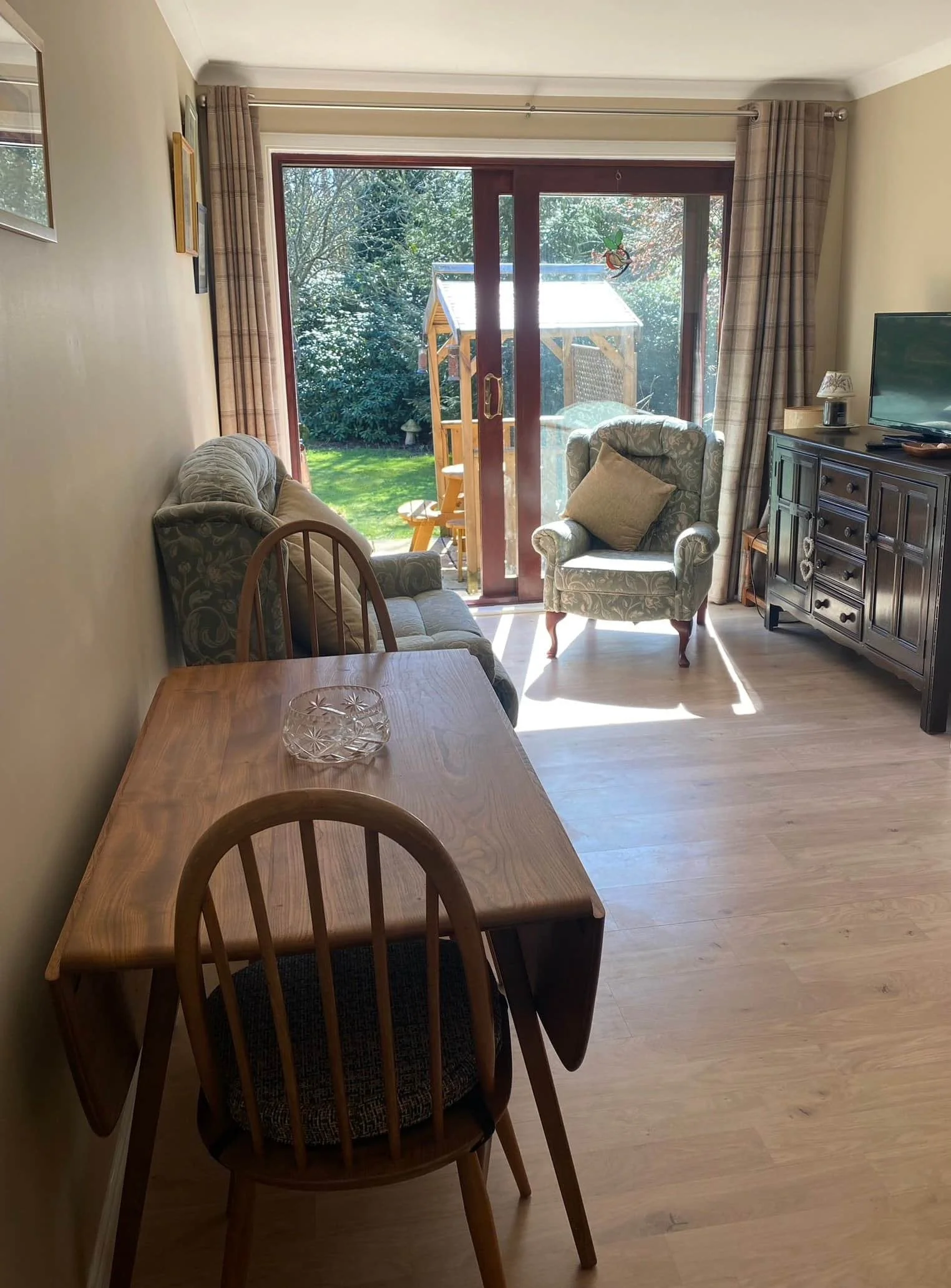 Living room with wooden dining table, chairs, patterned sofa, armchair with cushion, TV on a cabinet, large glass sliding door leading to a garden with a wooden playhouse.
