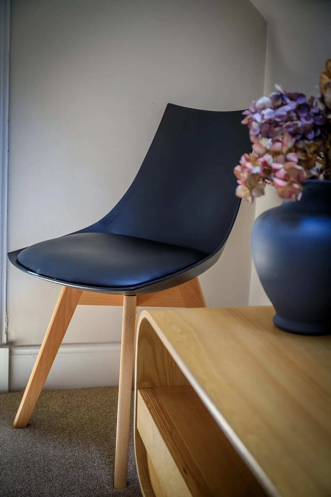 A black modern chair with wooden legs next to a wooden table with a dark vase of dried pink and purple flowers