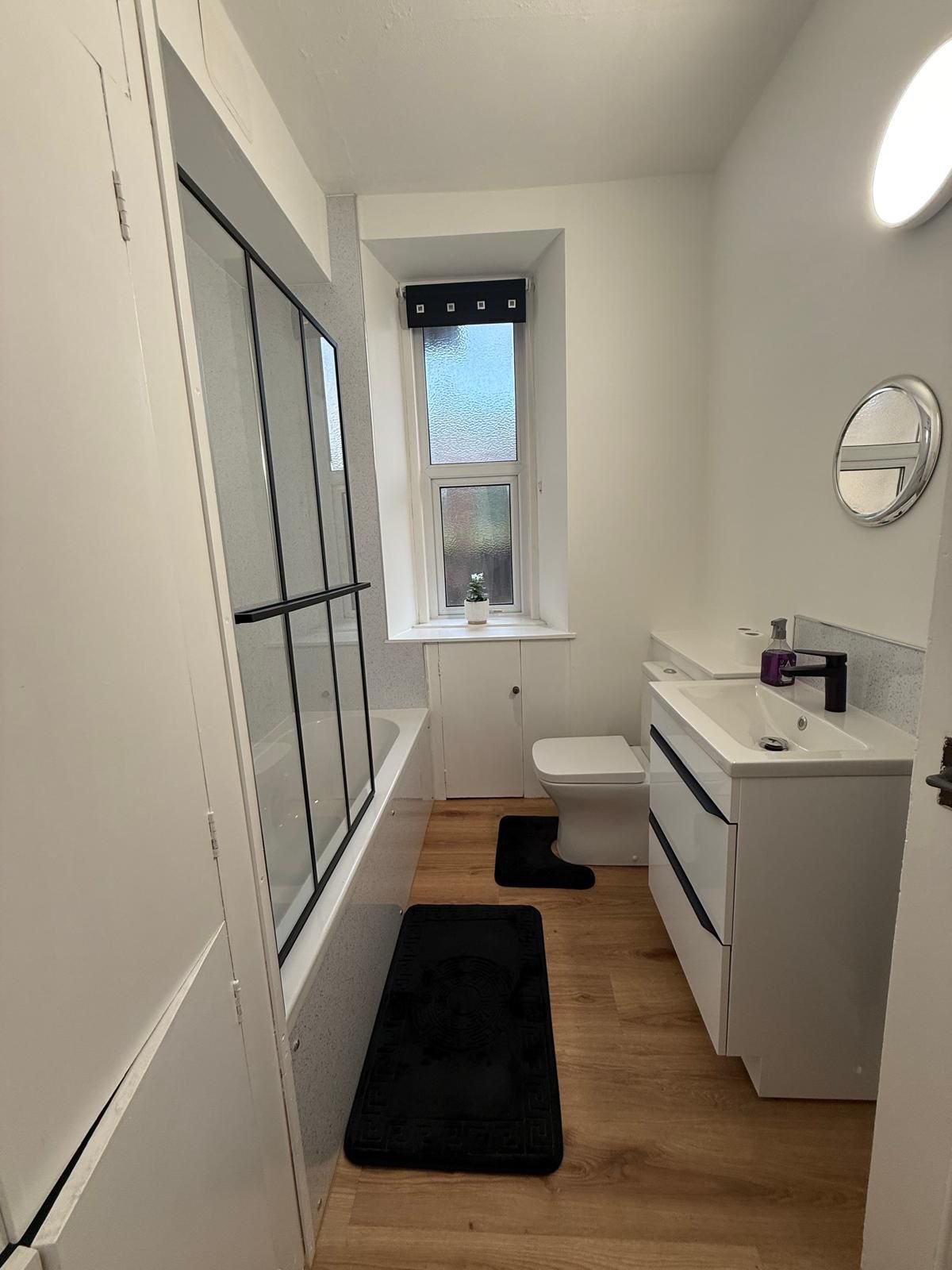 A small bathroom with a window, a white toilet, a white vanity with a black faucet, a round wall mirror, black bathroom rugs, and a bathtub with a black-framed glass shower door.