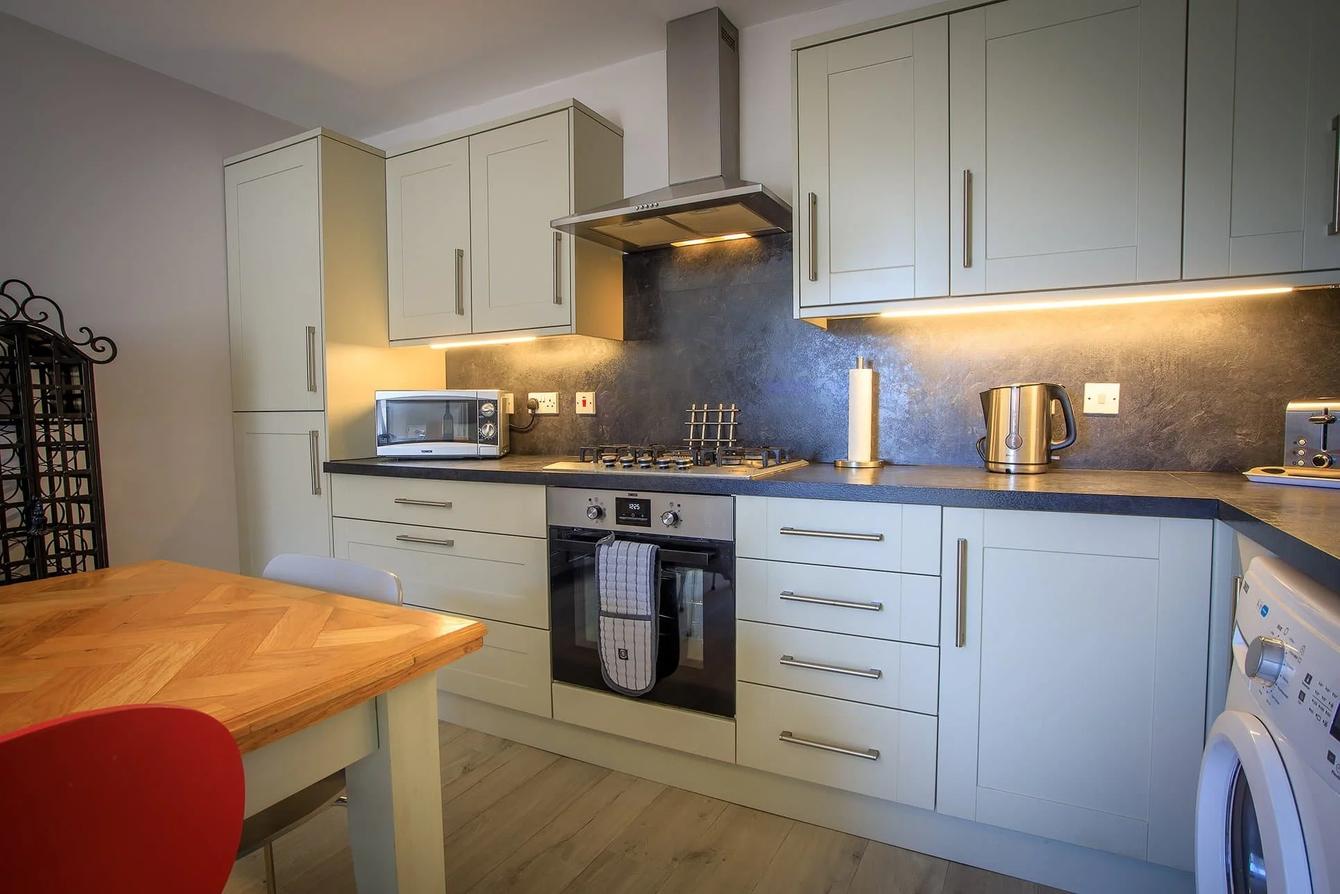 Modern kitchen with white cabinets, black countertops, stainless steel appliances, microwave, kettle, and washing machine. Dining table with red and white chairs.