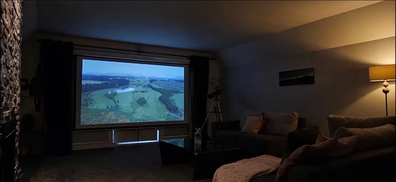 Living room with a large projection screen displaying a landscape view of a river and green fields, with couches, a lamp, and a telescope in the background.