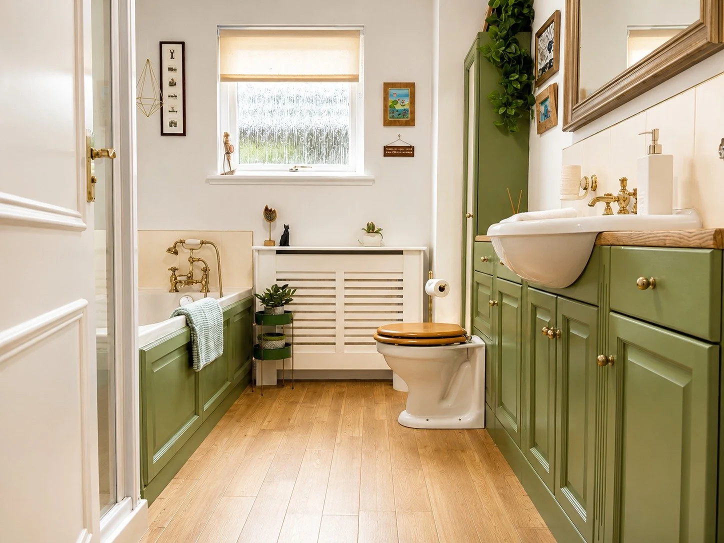 A cozy bathroom with green cabinetry, a white sink, and gold hardware. There is a bathtub with a green panel, a window with frosted glass, and various decorative items including plants and art on the walls. The floor is wooden, and there is a toilet 