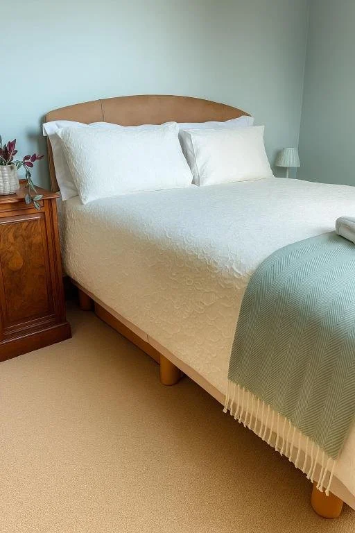 A neatly made bed with white bedding and pillows, a wooden nightstand with a vase of purple flowers, and a small lamp against a light-colored wall.