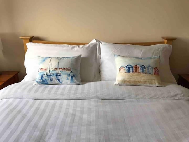 A neatly made bed with white linens, two large pillows, and two decorative pillows featuring beach and boat scenes, with wooden headboard and side tables.