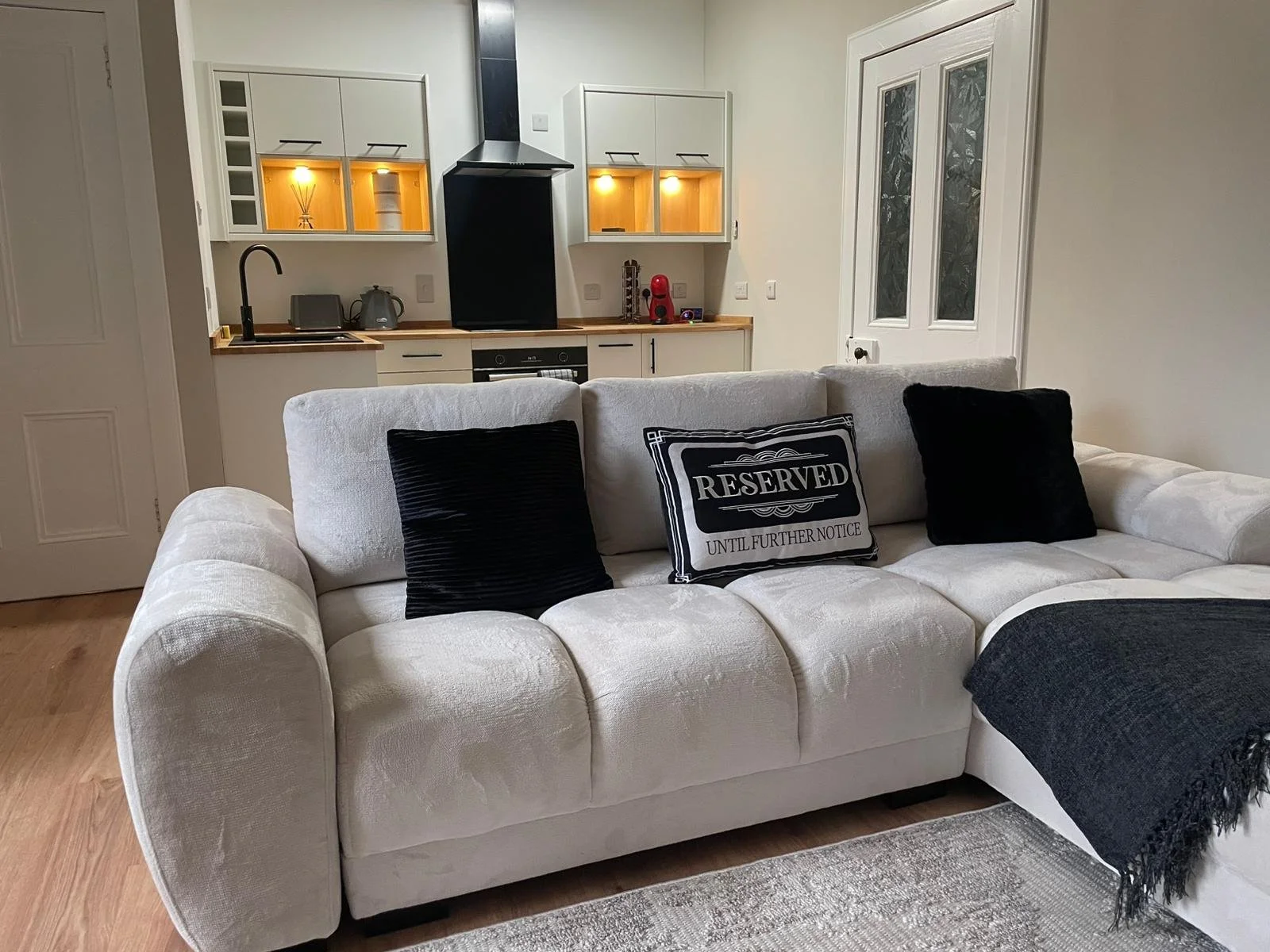 Living room with white sectional sofa, black and 'reserved' pillows, dark throw blanket, and open kitchen with white cabinets, wooden countertop, and small appliances in the background.