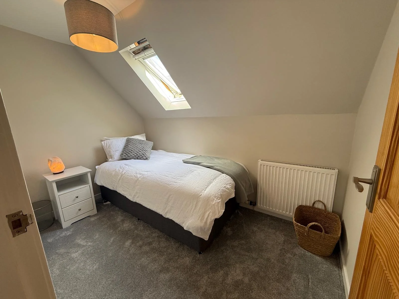A cozy bedroom with a bed, gray and white bedding, a small white nightstand with a salt lamp, a gray pillow, and a gray throw blanket. There is a skylight window on the slanted ceiling, a radiator on the wall, a wicker basket next to it, and a gray c