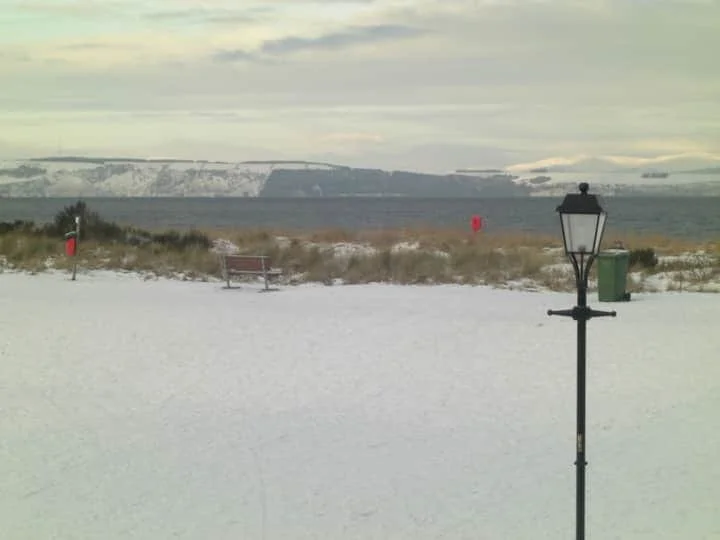 A snow-covered beach with a bench, a lamppost, and a green trash bin, with snow-covered hills in the background under a cloudy sky.