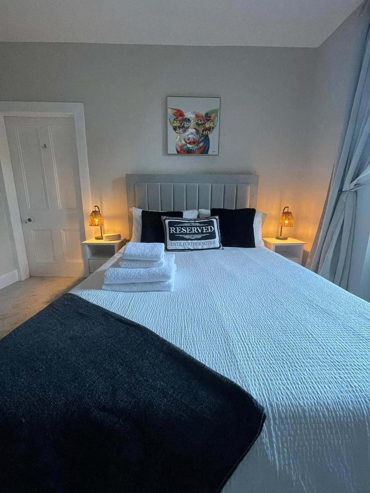 A neatly made bed in a bedroom with gray headboard, white bedding, and black pillows with a reservaed sign. Two bedside tables with lamps, a painting of a pig on the wall, and a door to the left. Towels are stacked on the bed.