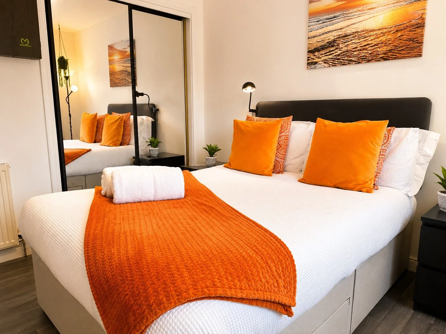 A neatly made bedroom with a bed that has white sheets, two white towels, and orange bedding. The bed has four orange pillows and two patterned pillows. There are small black nightstands on each side of the bed, each holding a small potted plant. A l