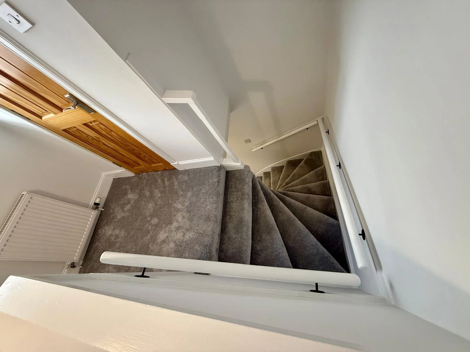 Indoor staircase with dark gray carpet, white walls, and a brown wooden door at the top, viewed from the bottom of the stairs.