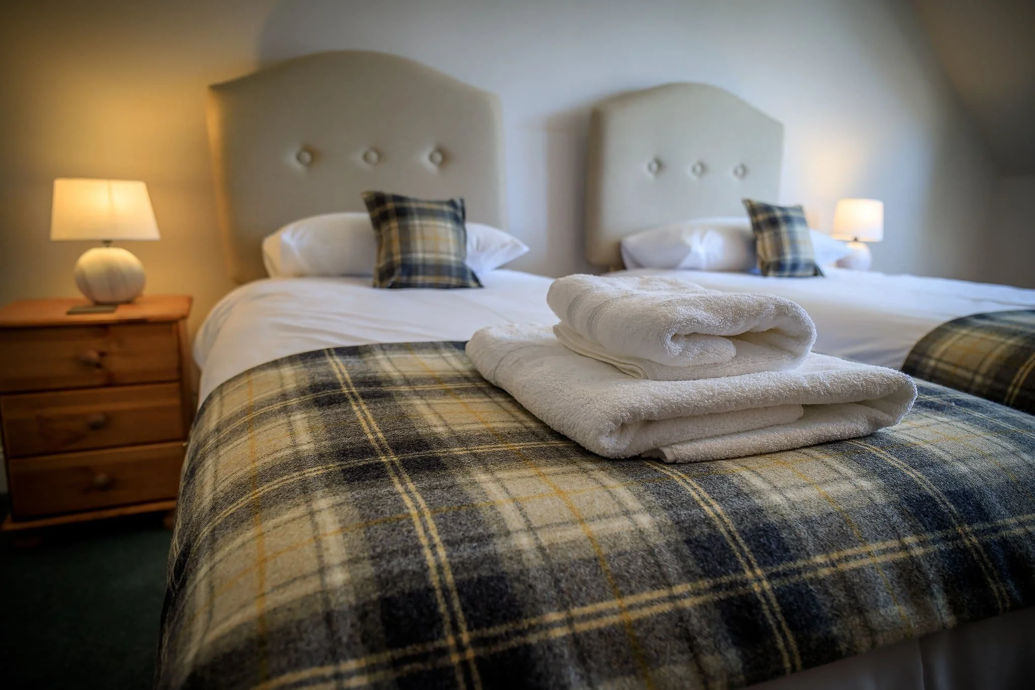 A neatly made bed with white sheets, plaid pillows and a matching plaid bedspread, with rolled white towels on top. The bed has two beige, tufted headboards and is flanked by wooden nightstands with table lamps, in a cozy bedroom setting.