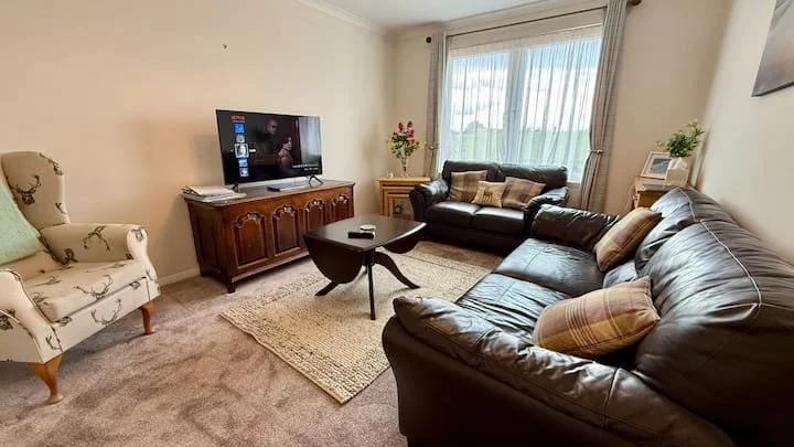 Living room with a TV on a wooden cabinet, a patterned armchair, two black leather sofas with pillows, a small coffee table, a large window with curtains, and a carpeted floor.